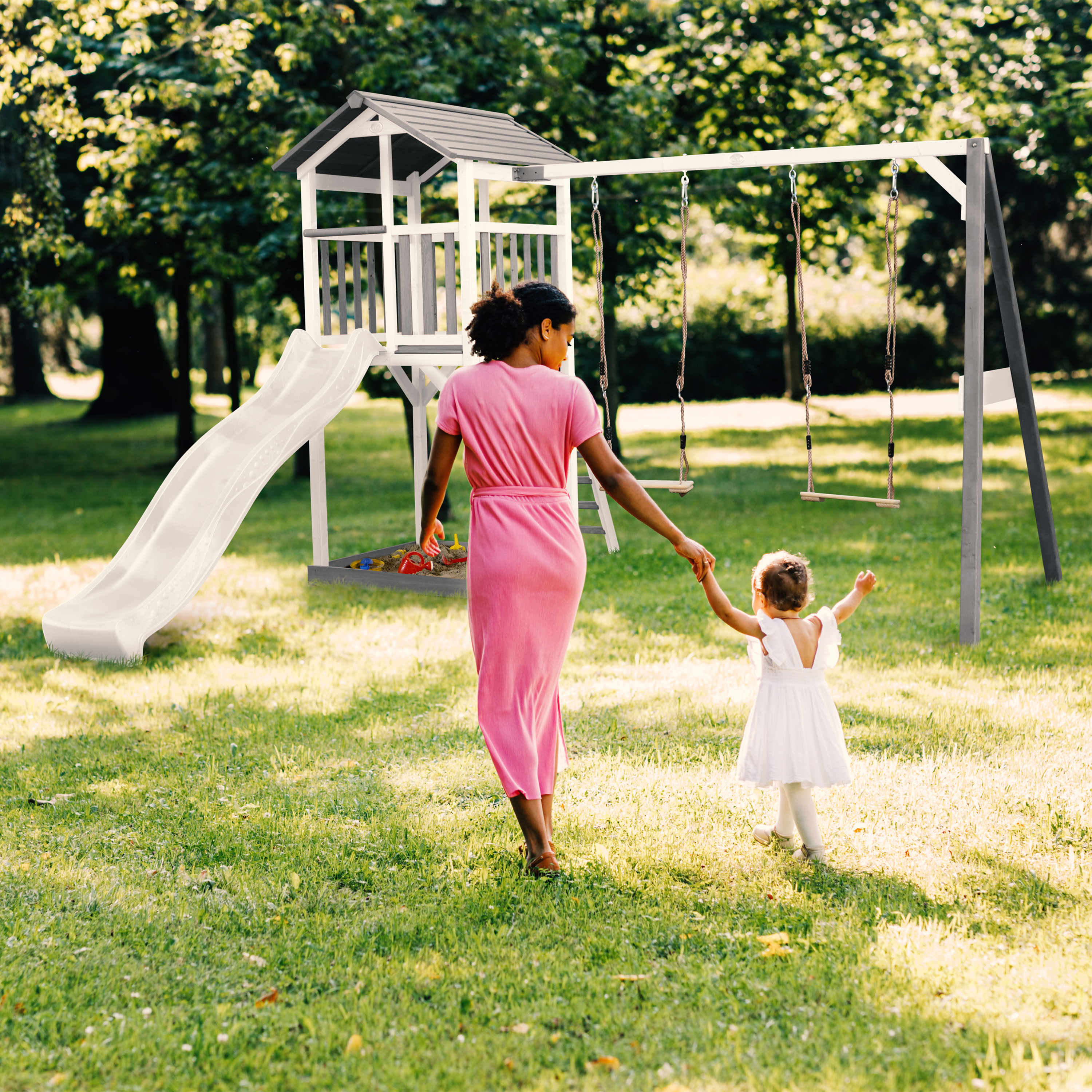 Frau und Kind gehen zu Spielplatz mit Rutsche und Schaukeln im Gras.