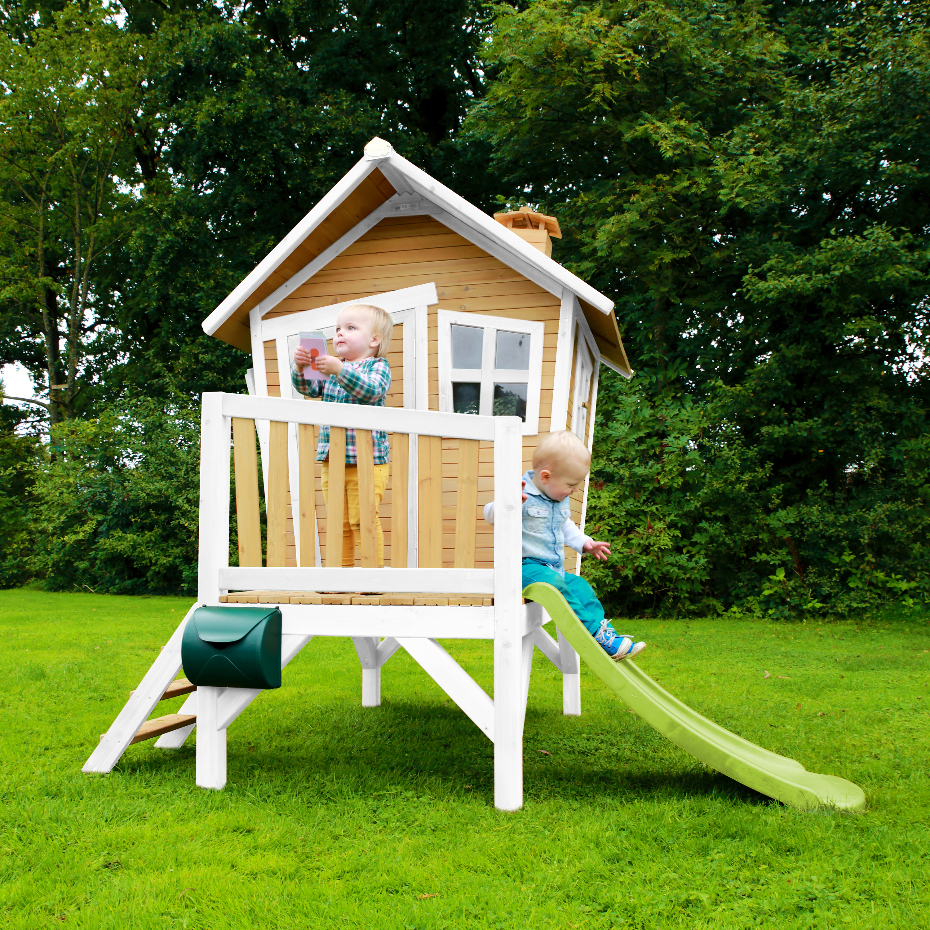 Zwei Kinder spielen auf einem Holzspielhaus mit Rutsche und Briefkasten im Garten.