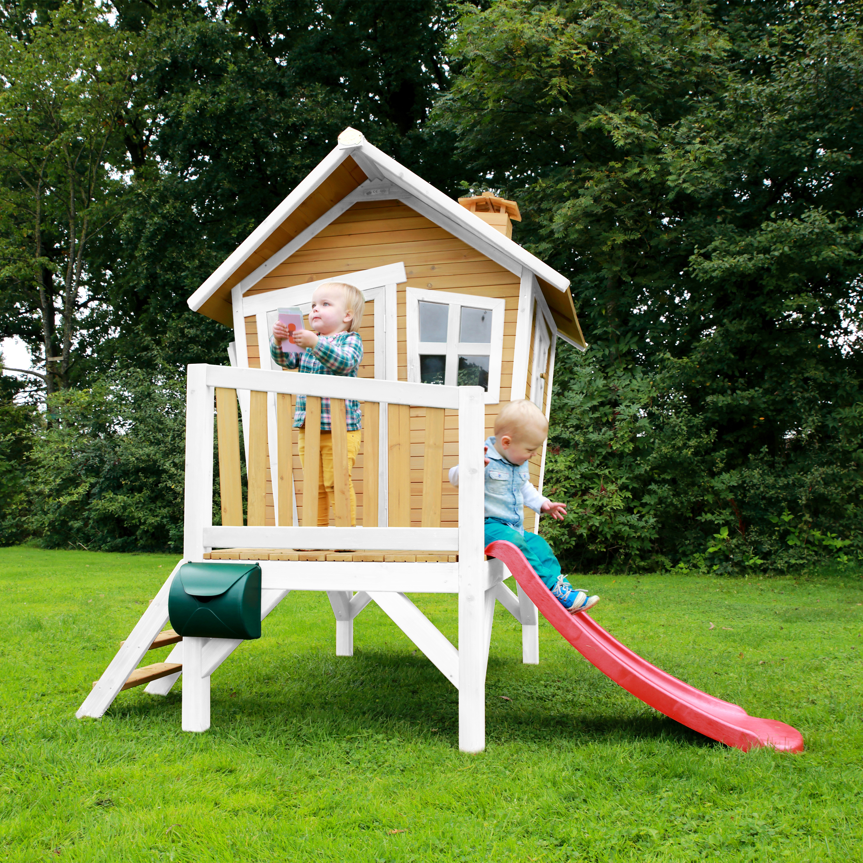Zwei kleine Kinder spielen auf einem Holzhaus mit Rutsche im Garten.