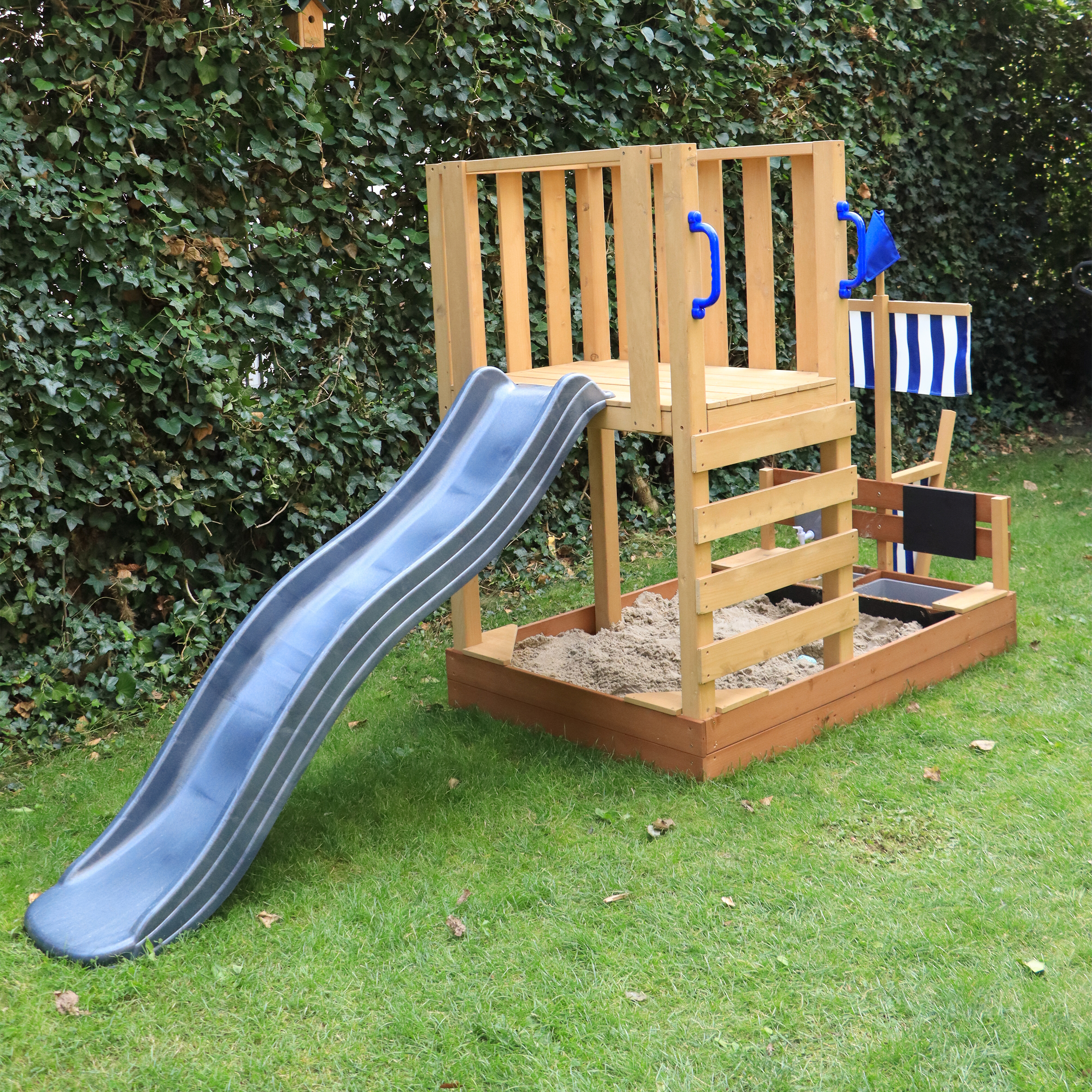 Holzspielturm mit Rutsche, Sandkasten und blau-weißer Fahne im Garten.