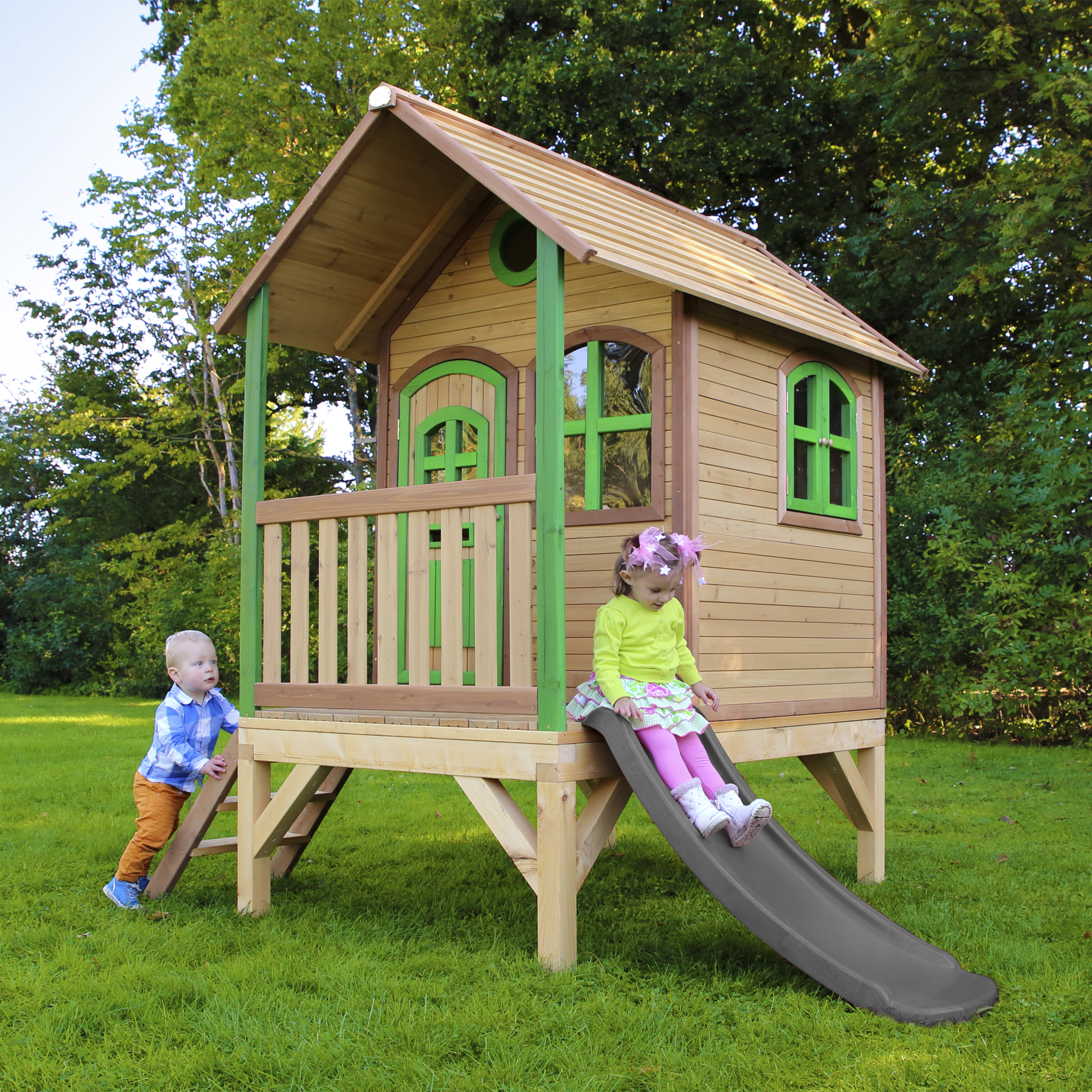 Zwei Kinder spielen an einem Holzspielhaus mit Rutsche im Garten.