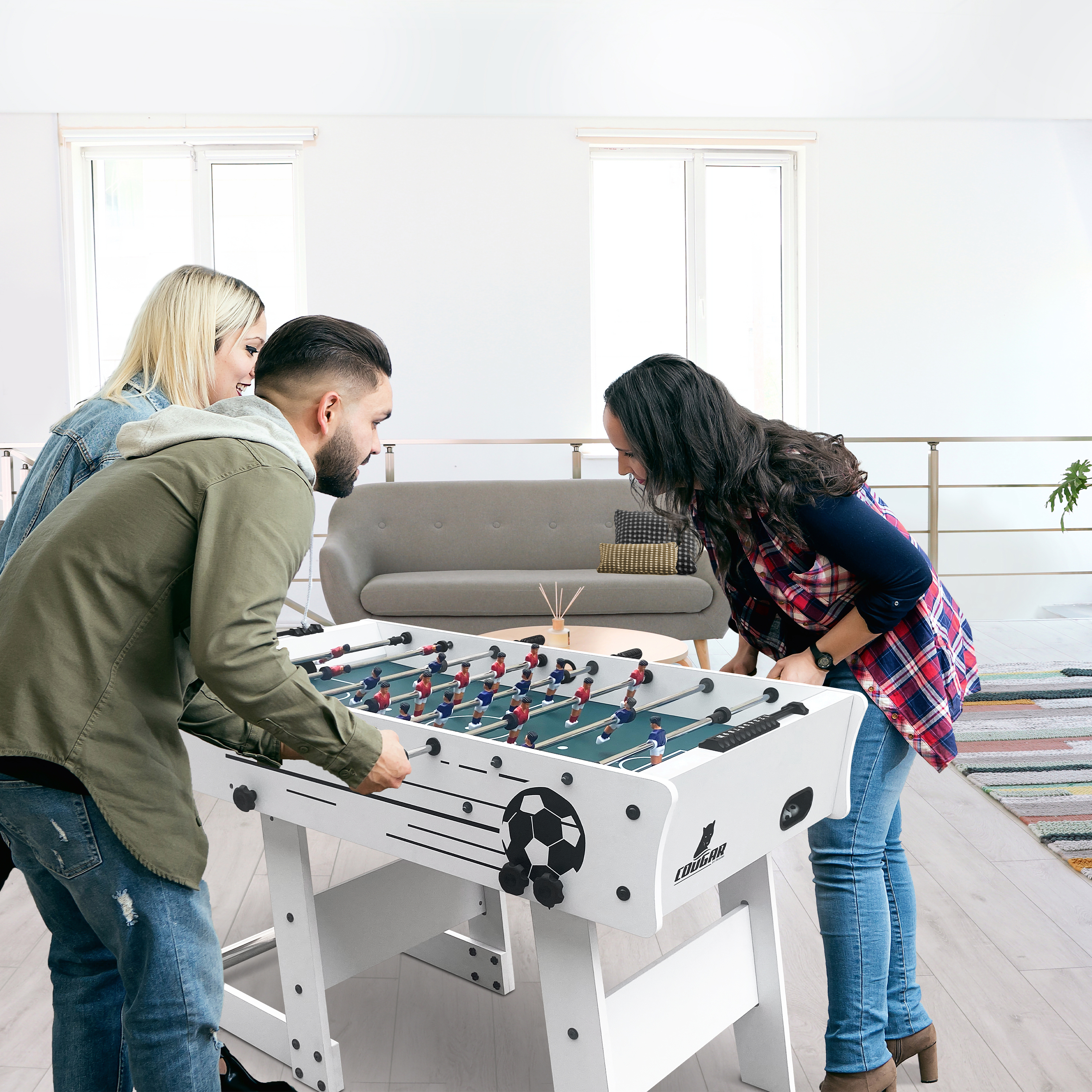 Vier Personen spielen Tischfußball in einem hellen Raum mit Sofa und Teppich.