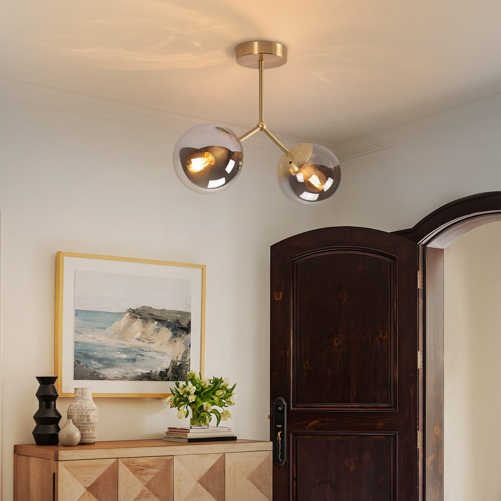 Lampada da soffitto dorata con due globi di vetro sopra una cassettiera con arte e vasi.