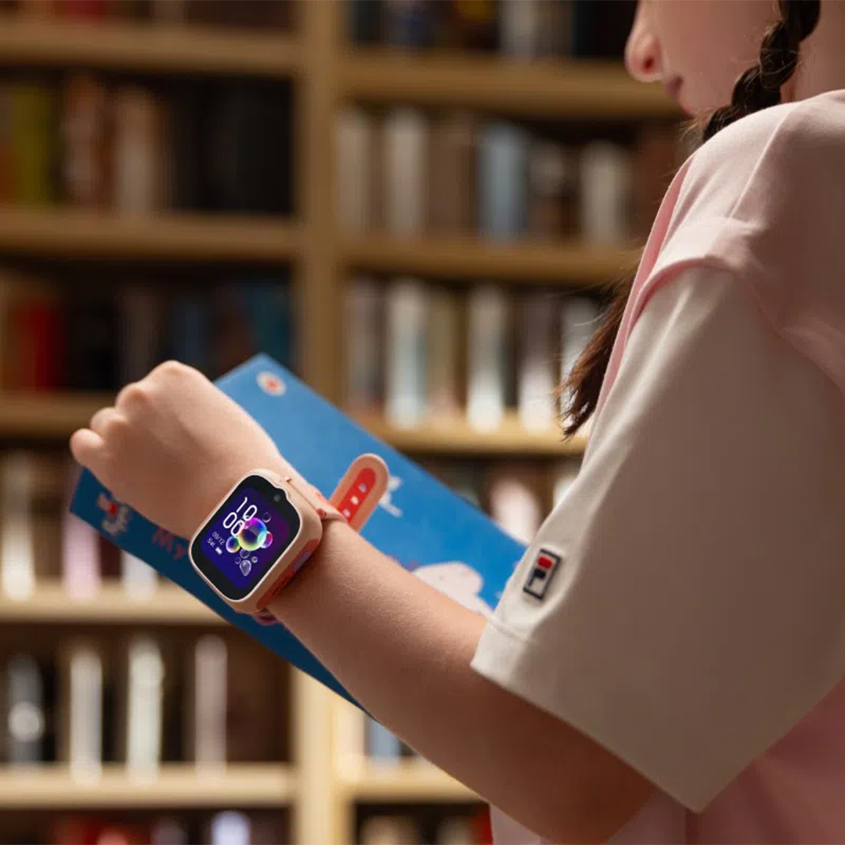 Un niño lleva un smartwatch mientras sostiene un libro frente a estanterías.