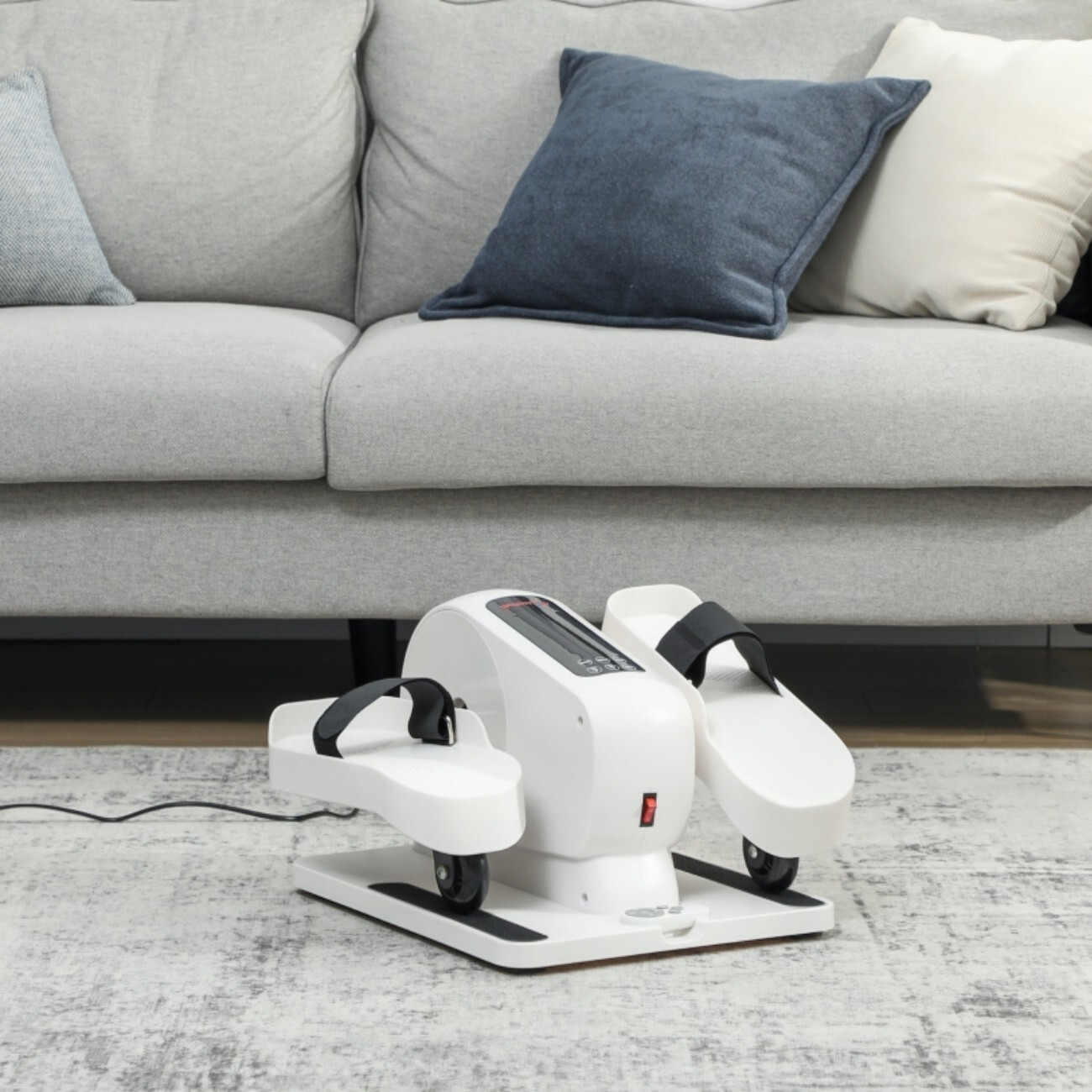 Biała domowa maszyna do ćwiczeń z paskami na stopy stoi na dywanie przed sofą.