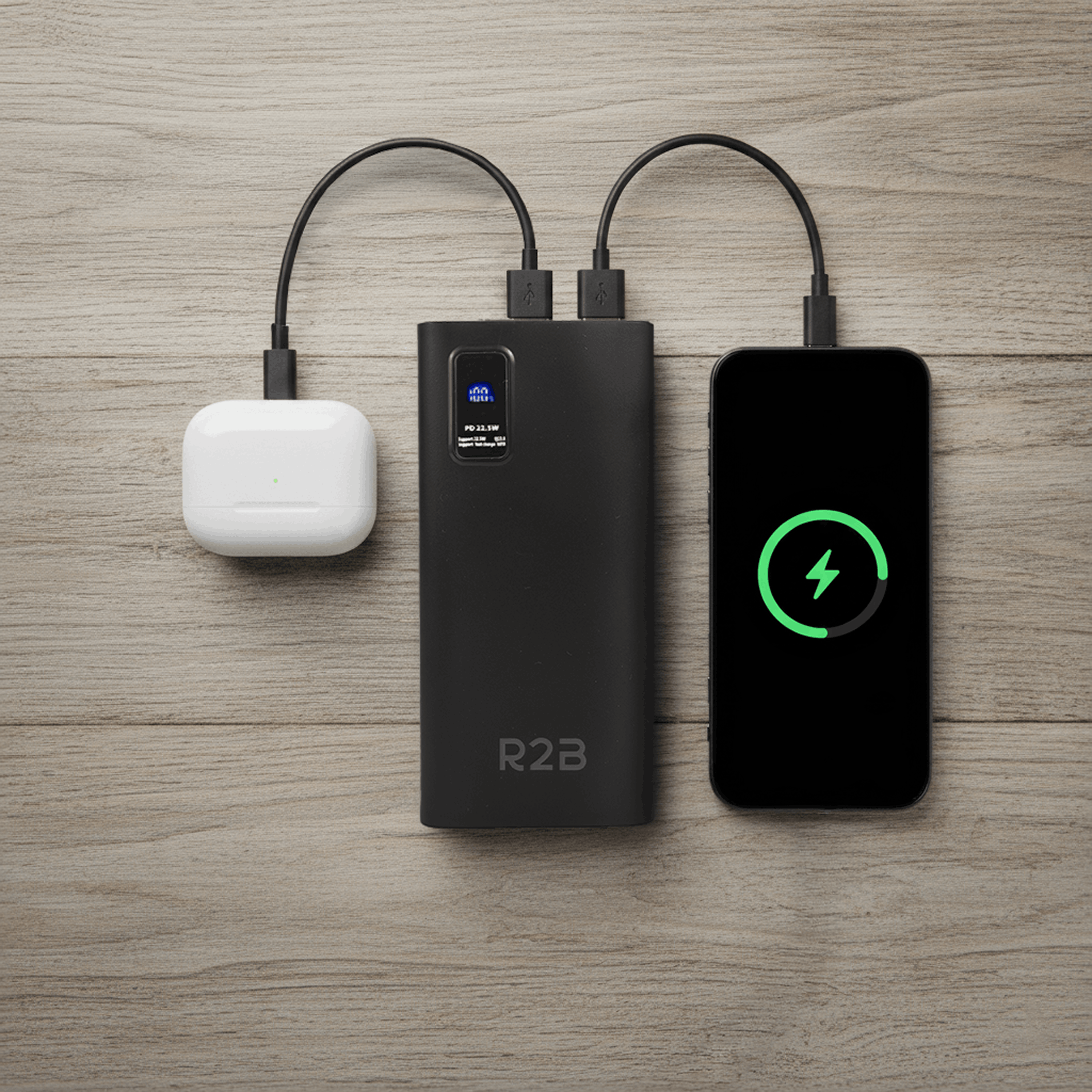Black power bank charges white earbuds and a smartphone on a wooden table.