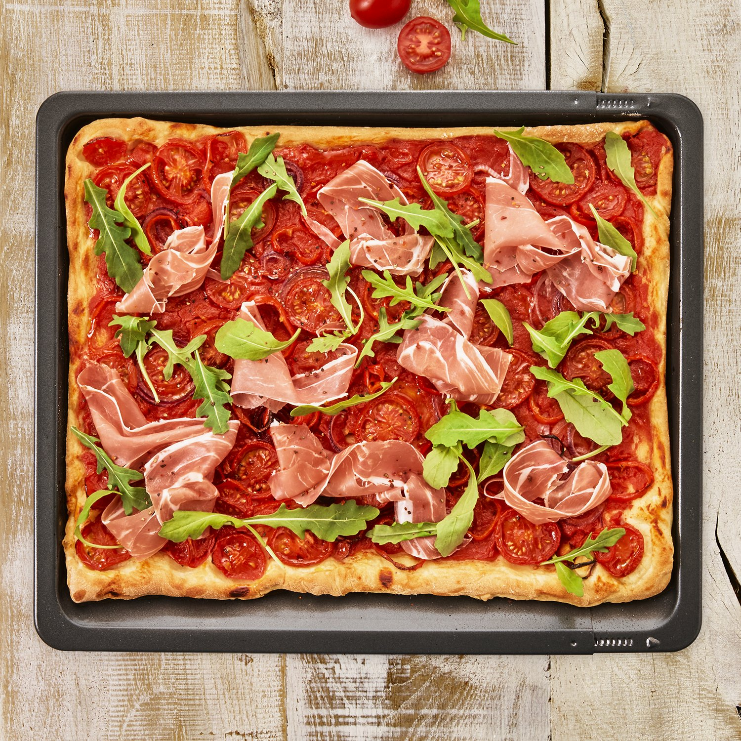 Rechteckige Pizza auf einem Backblech mit Tomaten, Prosciutto und Rucola.