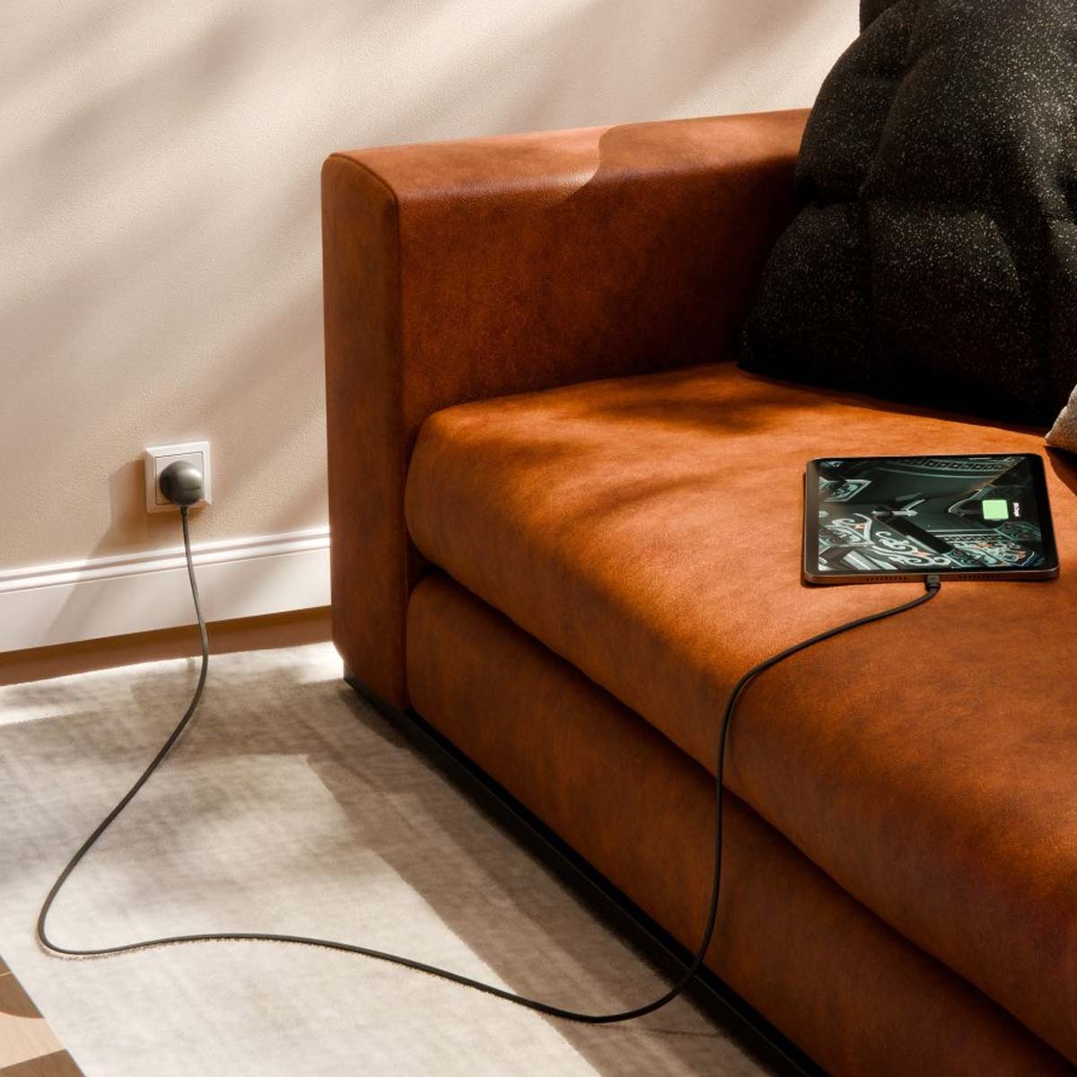 Un tablet si carica su un divano marrone, collegato a una presa a muro tramite cavo nero.