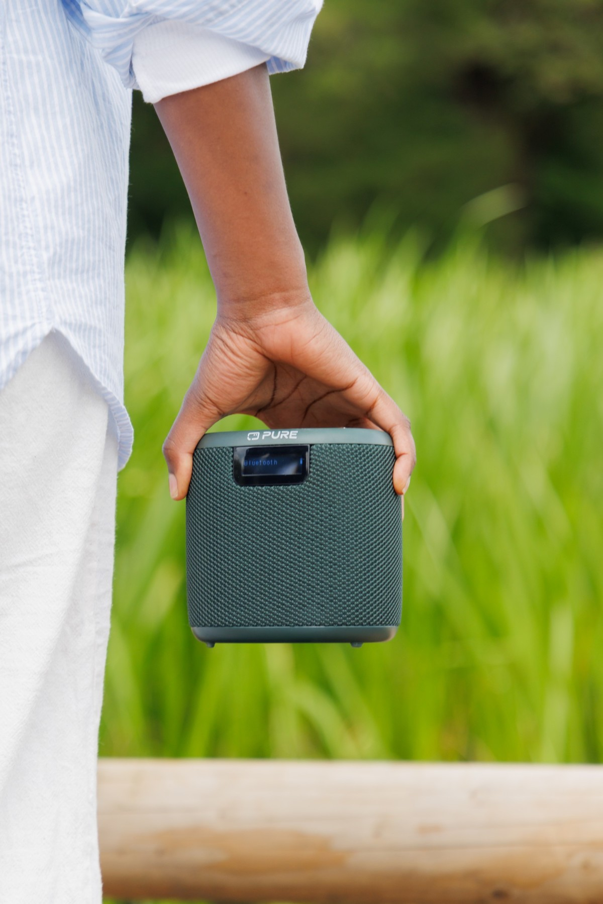 Une main tient un haut-parleur vert avec « Bluetooth » à l'écran, devant de l'herbe verte.