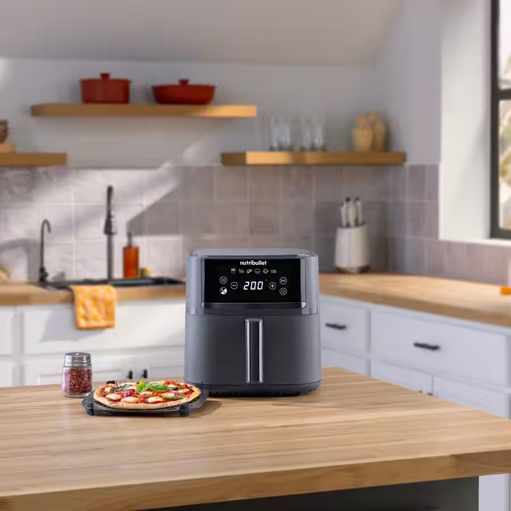 Ein dunkler Airfryer steht auf einer Holzoberfläche neben einer kleinen Pizza.