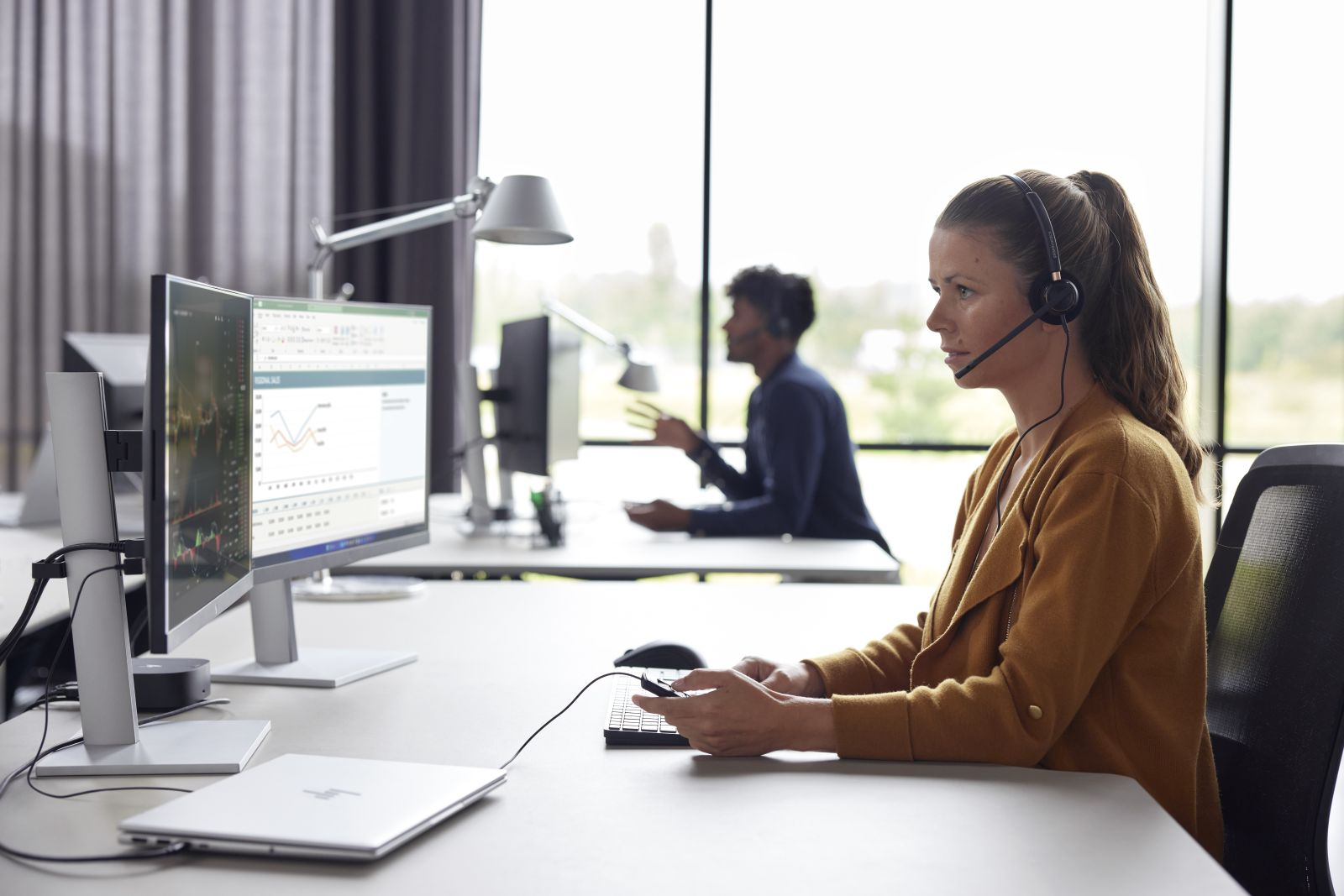Frau mit Headset am Schreibtisch vor zwei Monitoren, Mann im Hintergrund.