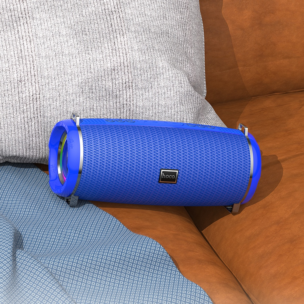 Un altavoz Bluetooth azul con detalles plateados descansa sobre un sofá junto a cojines.