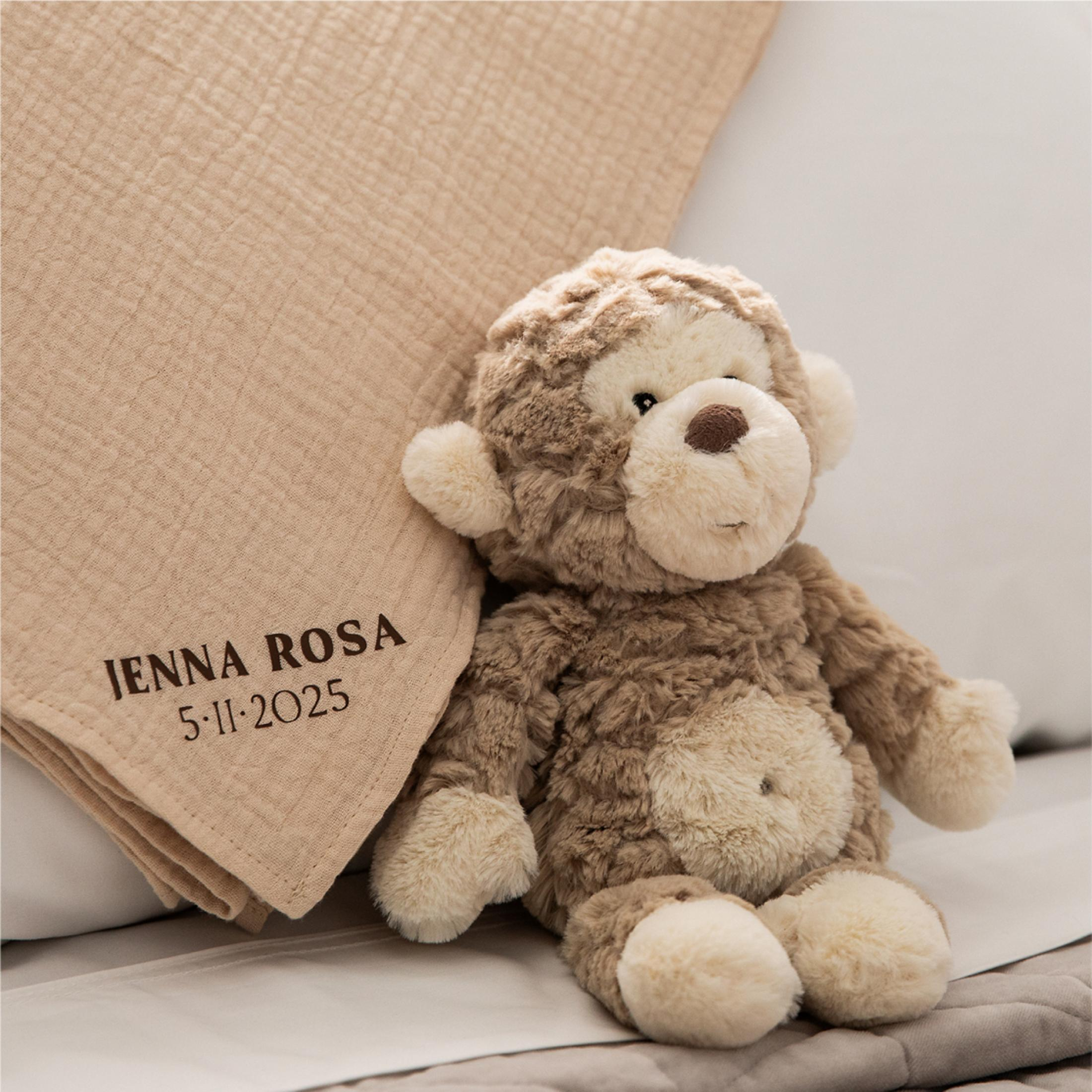 Ein brauner Teddybär sitzt neben einer beige Decke mit Namen und Datum.
