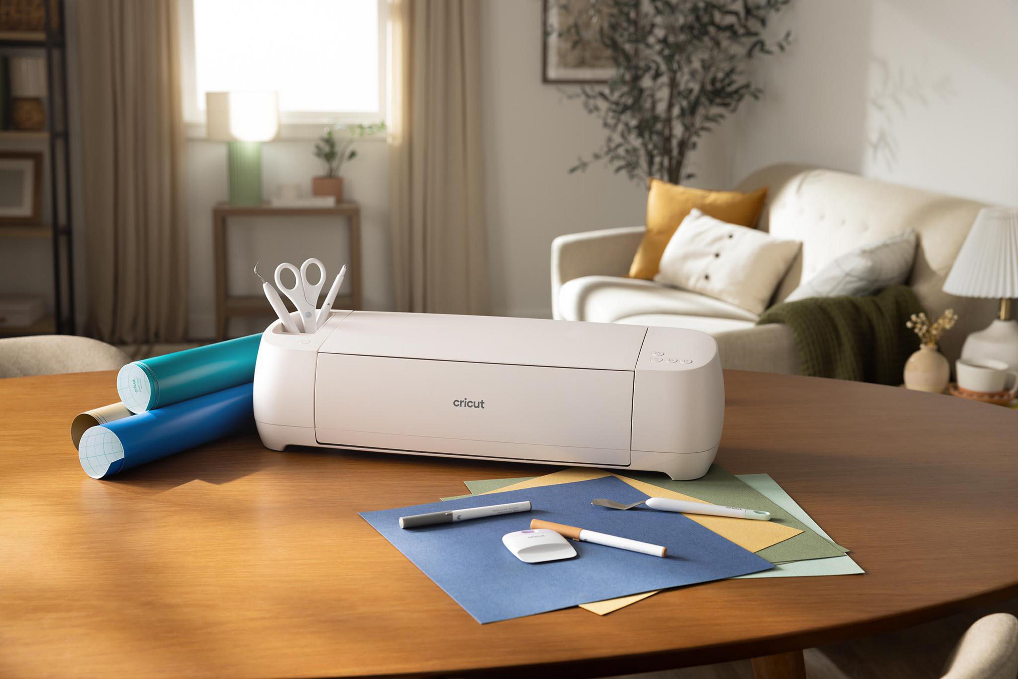 Ein weißer Cricut-Schneideplotter mit Bastelwerkzeugen und Papierrollen auf einem Holztisch.