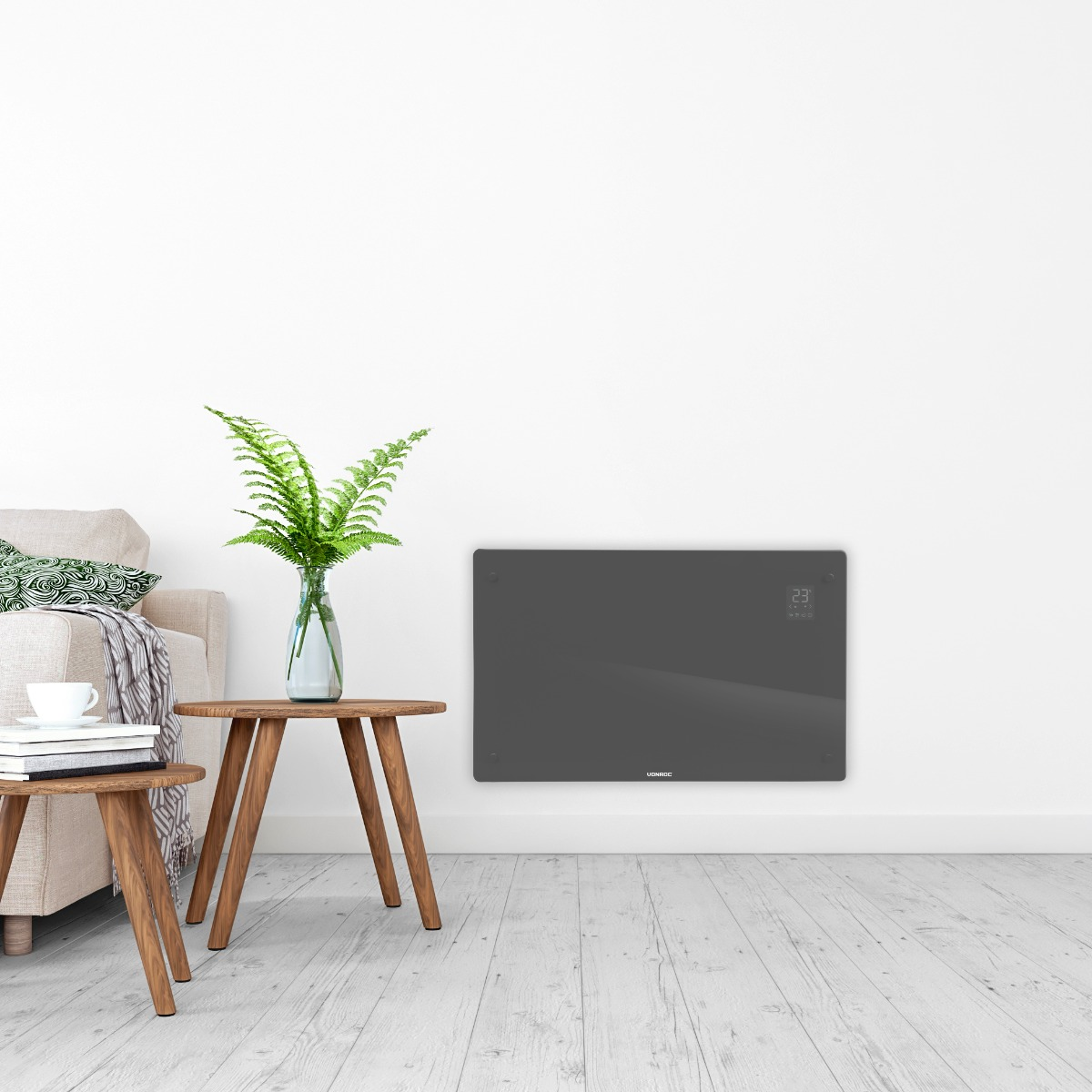 A gray electric heater with a digital display hangs on a white wall above a sofa and wooden tables.