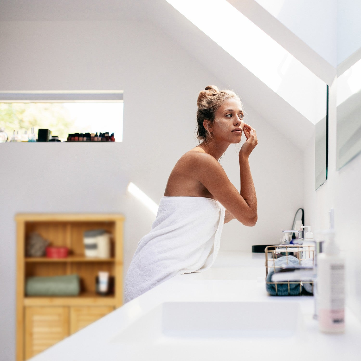 Mujer con mascarilla en un baño, aplicándose crema en la mejilla con el dedo.