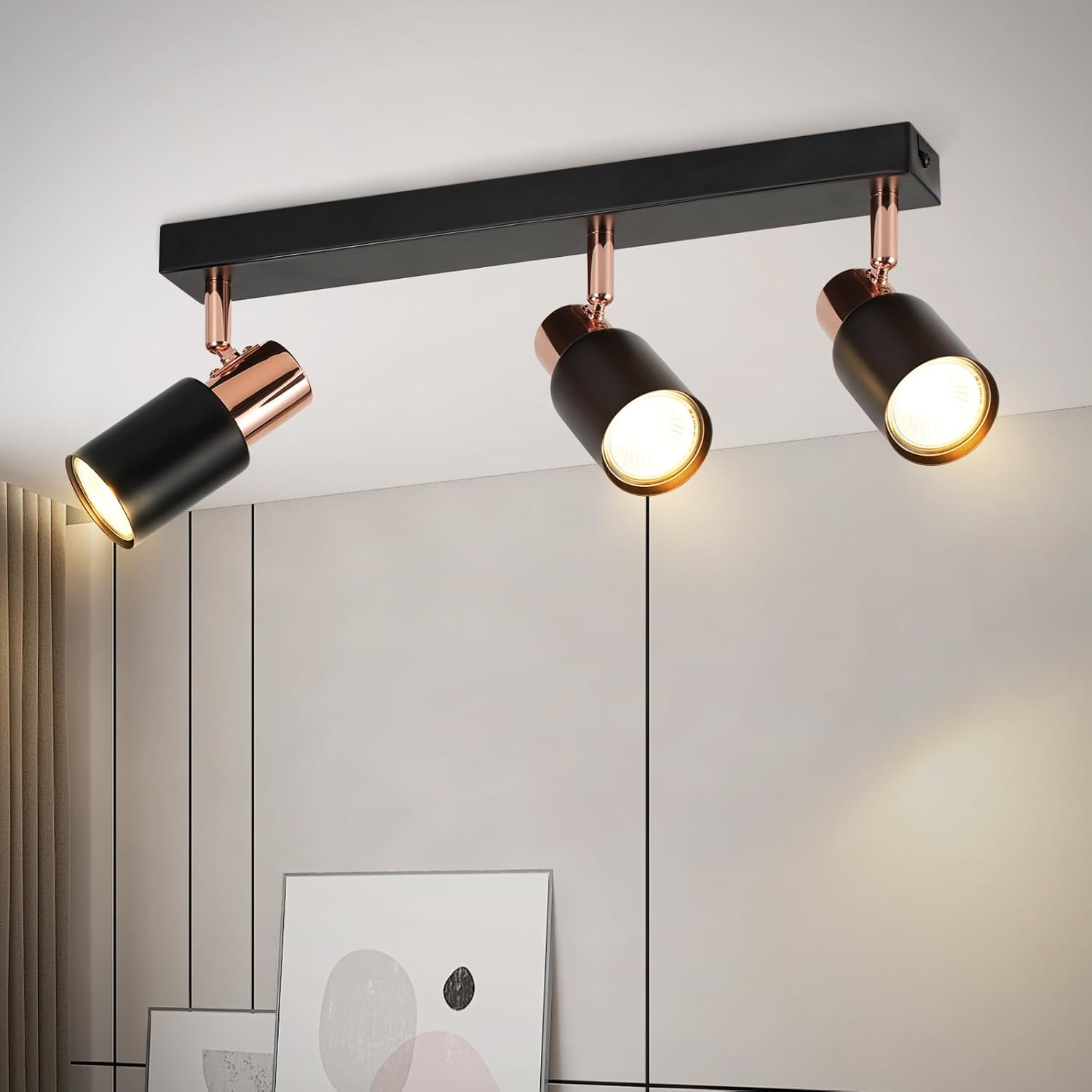 Lampada da soffitto nera e rame con tre faretti orientabili sopra quadri incorniciati.