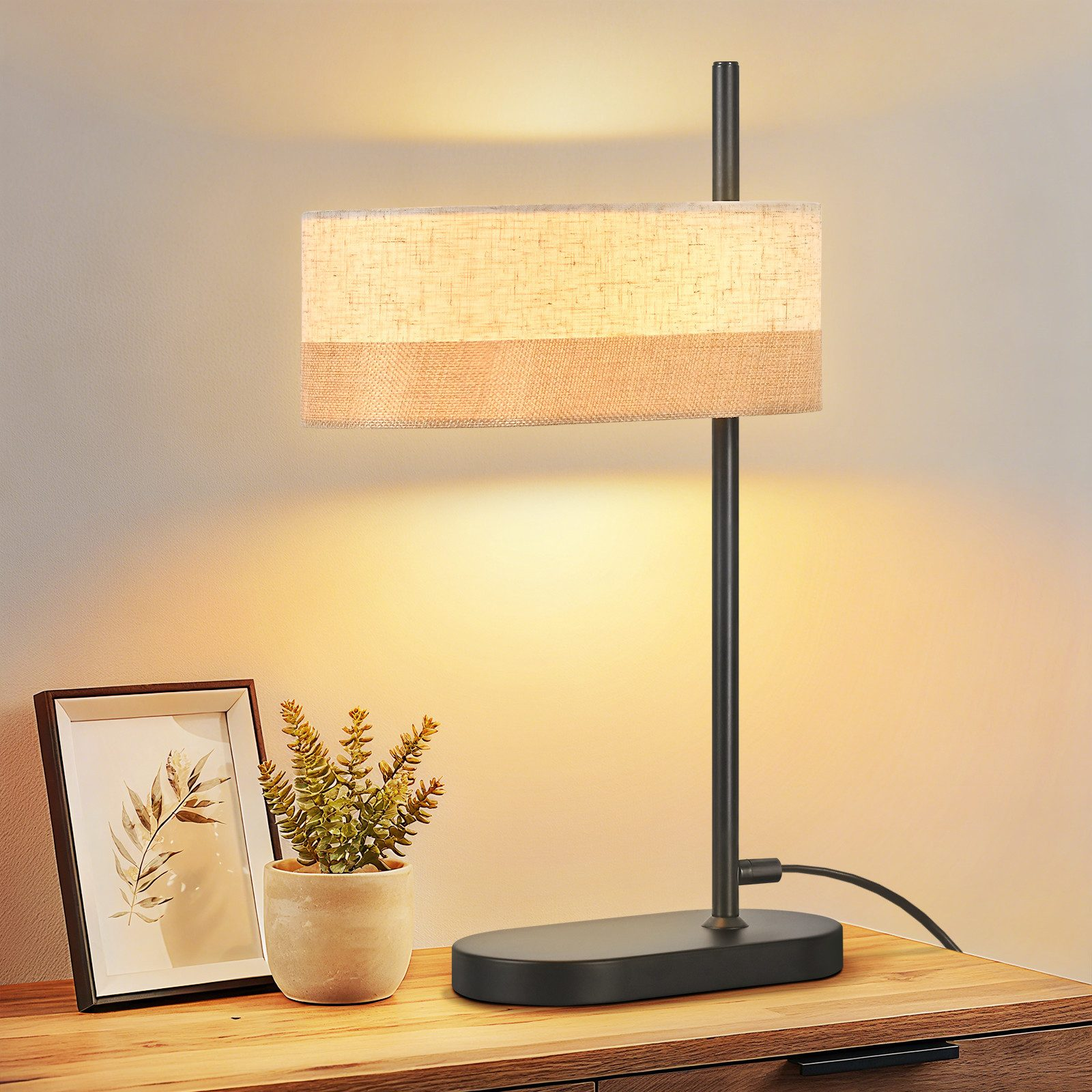 Una lampada rettangolare con paralume chiaro è su un comodino accanto a un quadro e una pianta.