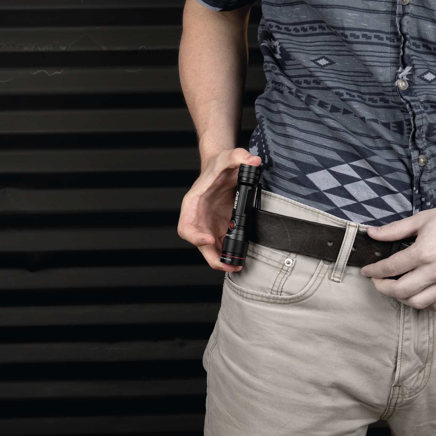 Dłoń trzyma czarną latarkę z czerwonymi akcentami obok jasnych dżinsów.