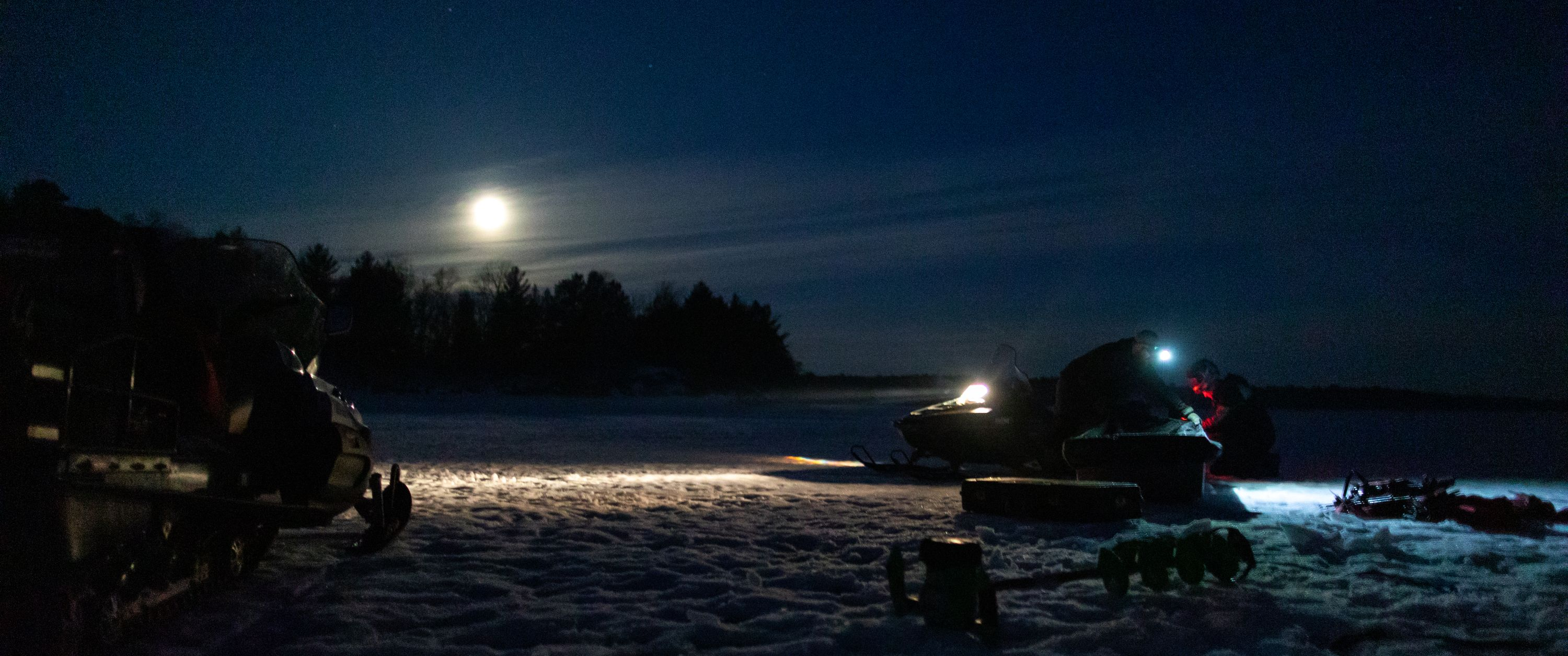 Schneemobile und Angler bei Nacht unter Vollmond auf gefrorenem See.