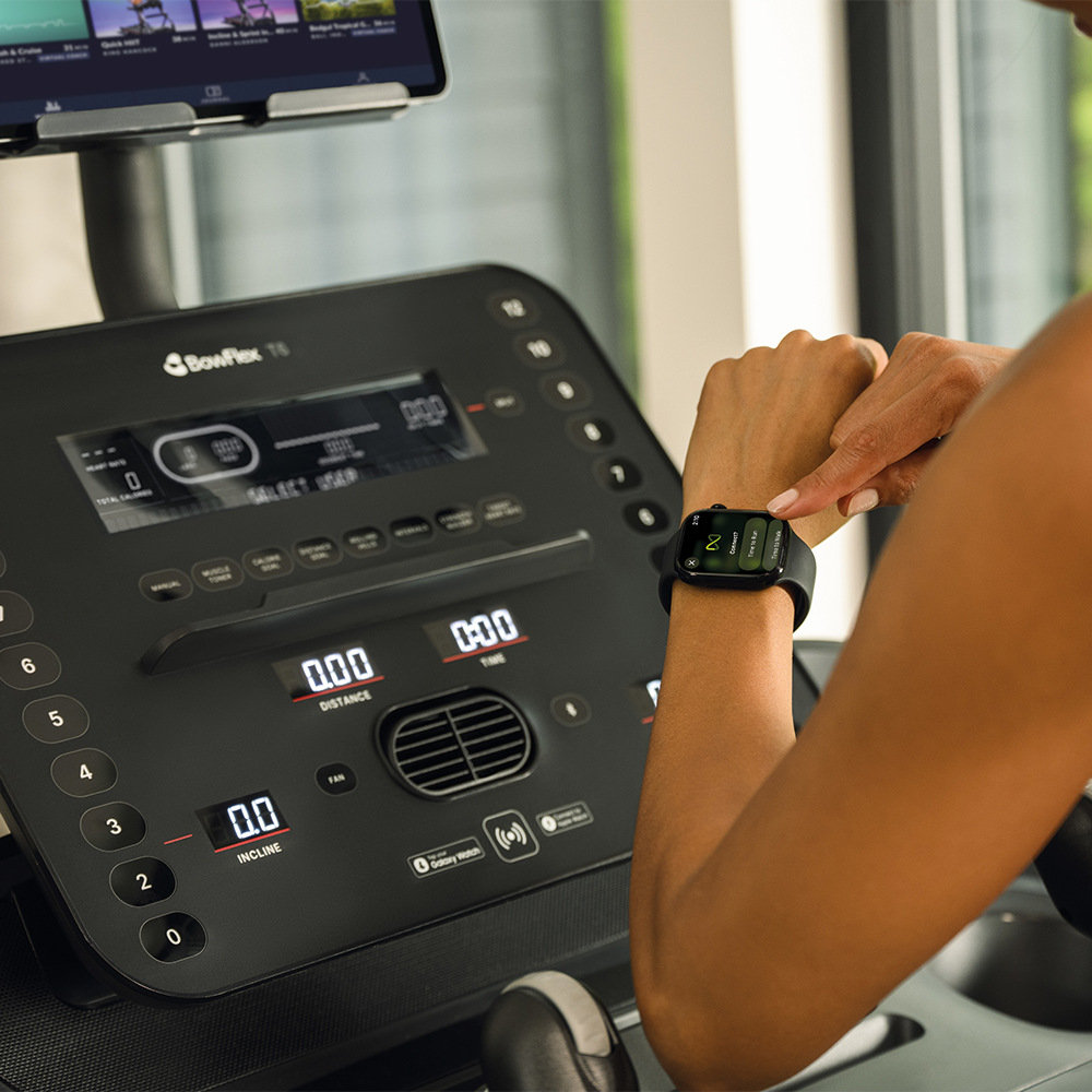 A woman checks her smartwatch display on a treadmill showing distance and incline stats.