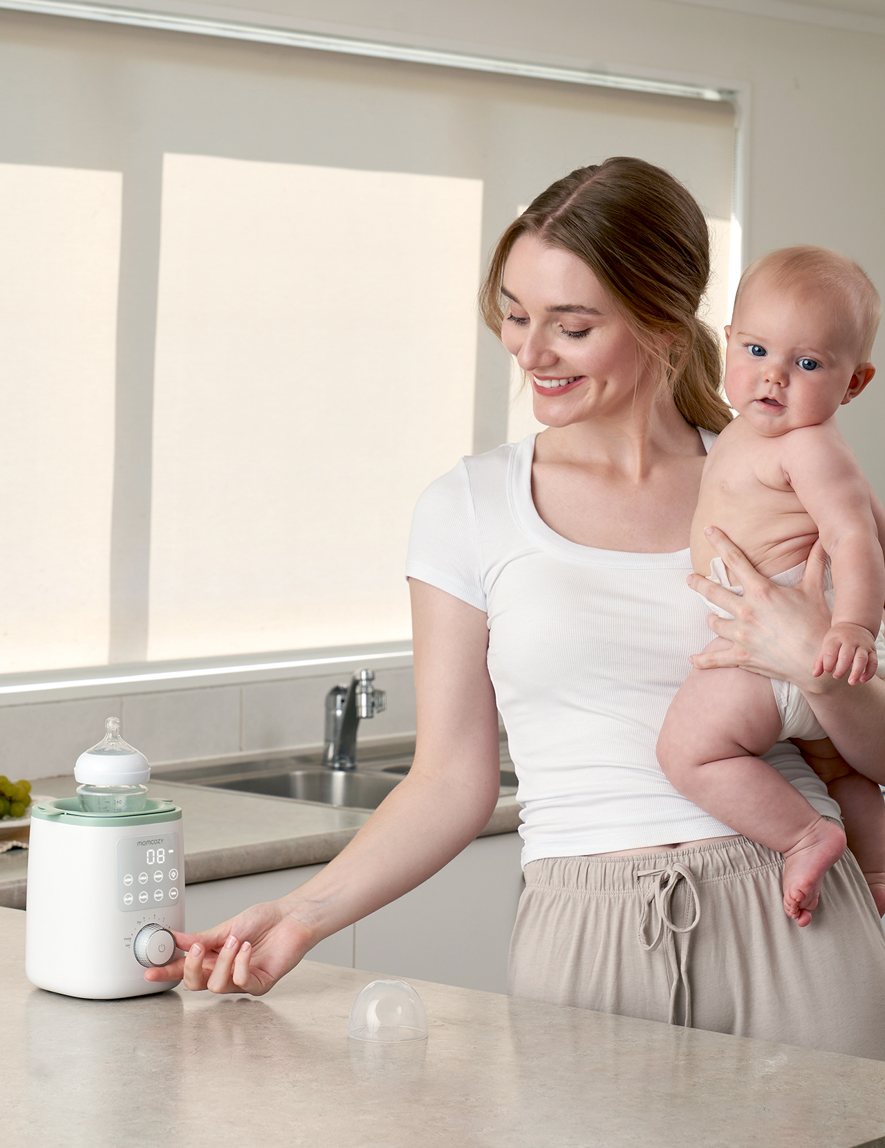 Eine Frau hält ein Baby und bedient einen Flaschenwärmer in einer Küche.