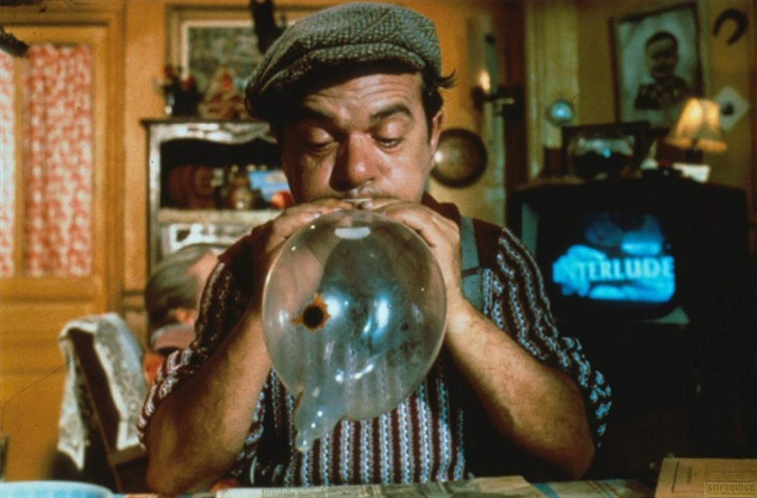 Un uomo con coppola gonfia una bolla trasparente, con un programma TV sullo sfondo.