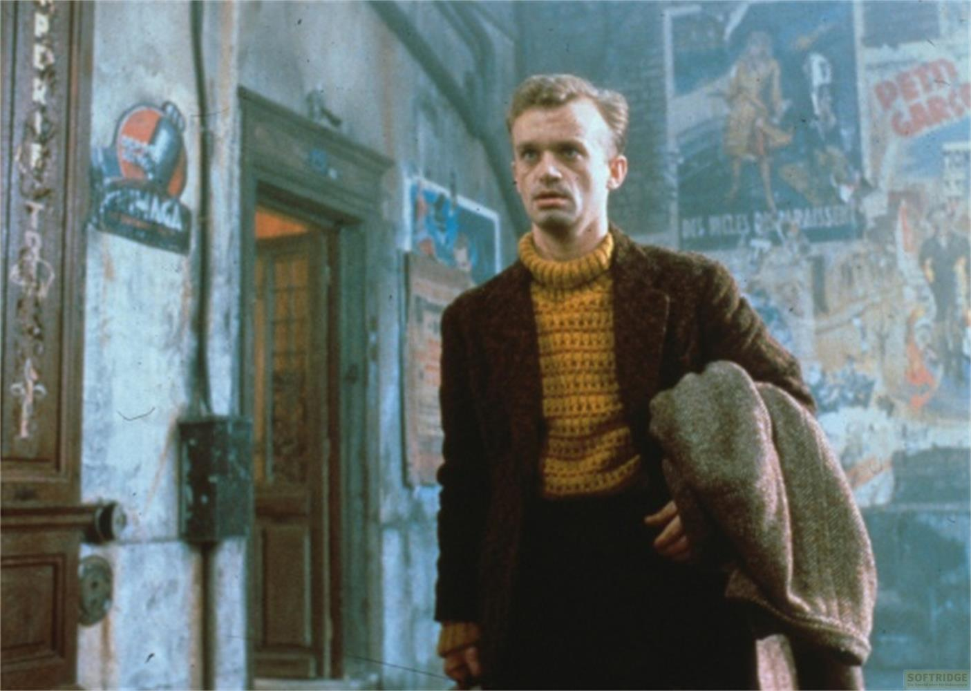 Un uomo con maglione giallo e cappotto marrone si trova davanti a un muro coperto di poster.