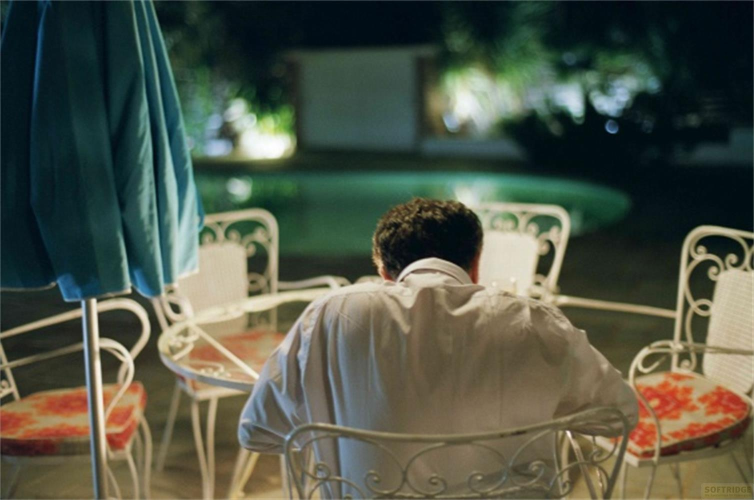 Uomo seduto a un tavolo all'aperto, con piscina e alberi sullo sfondo.
