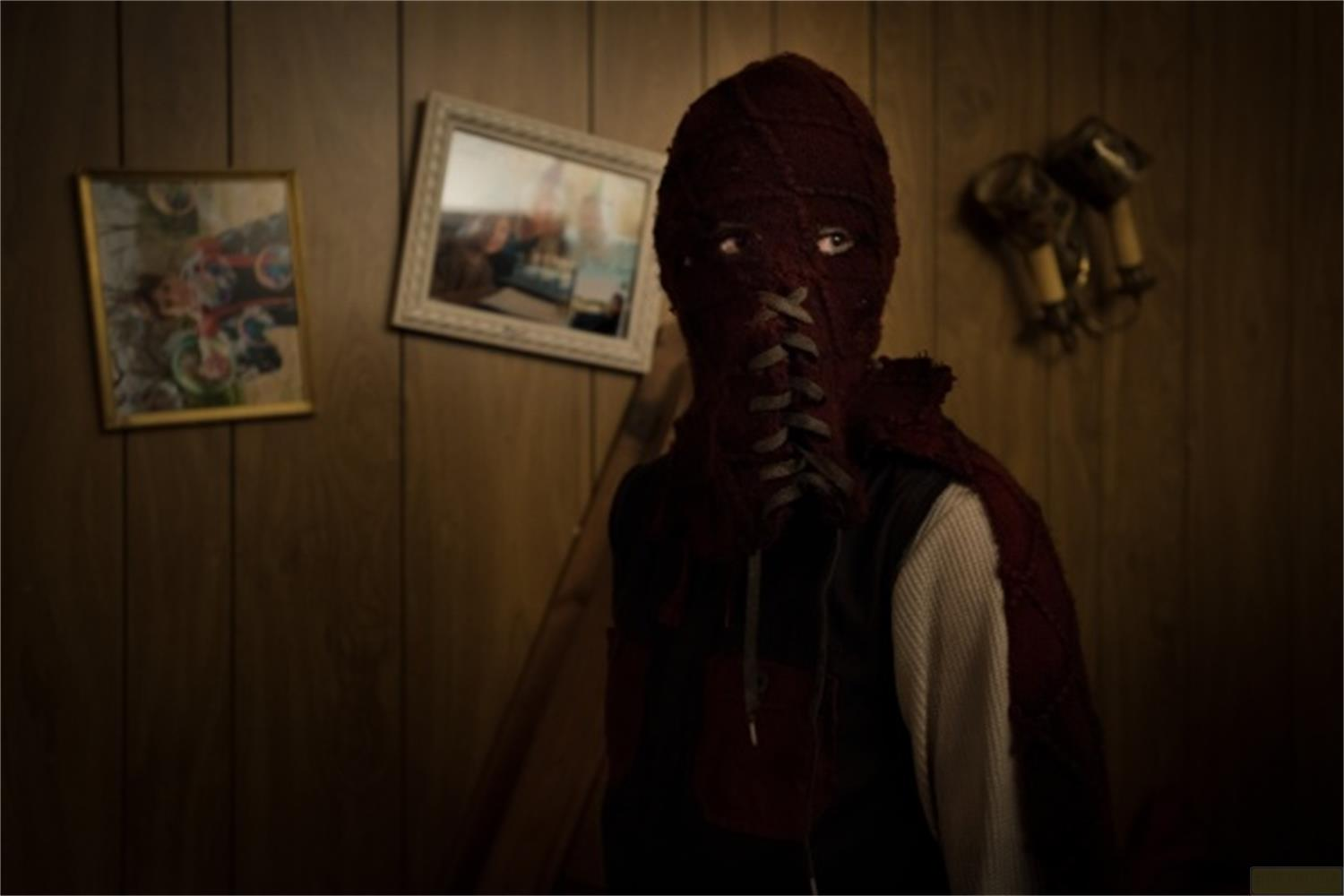 Una persona con maschera di lana rossa e lacci sul viso, davanti a una parete di legno.
