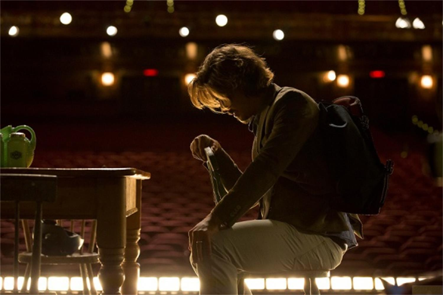 Un homme assis sur un tabouret, regardant vers le bas, tenant une bouteille. Sièges de théâtre.