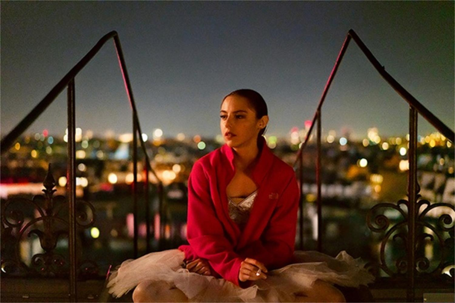 Una giovane donna in abiti da ballo siede all'aperto di notte con luci della città sullo sfondo.
