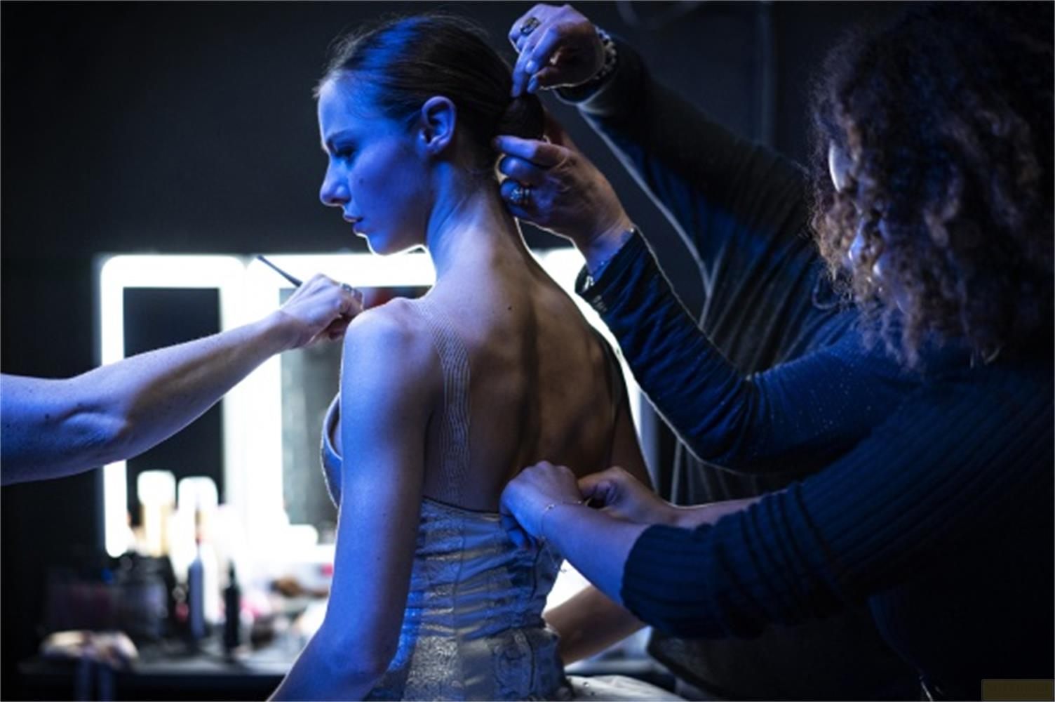 Ballerina pettinata da mani dietro le quinte, illuminata da luce blu.
