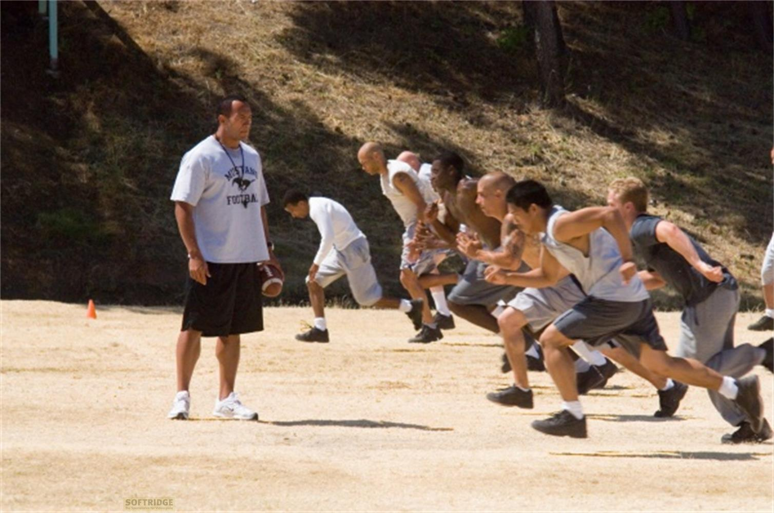 Ein Trainer mit Ball steht vor einer Gruppe von Football-Spielern in Startposition.