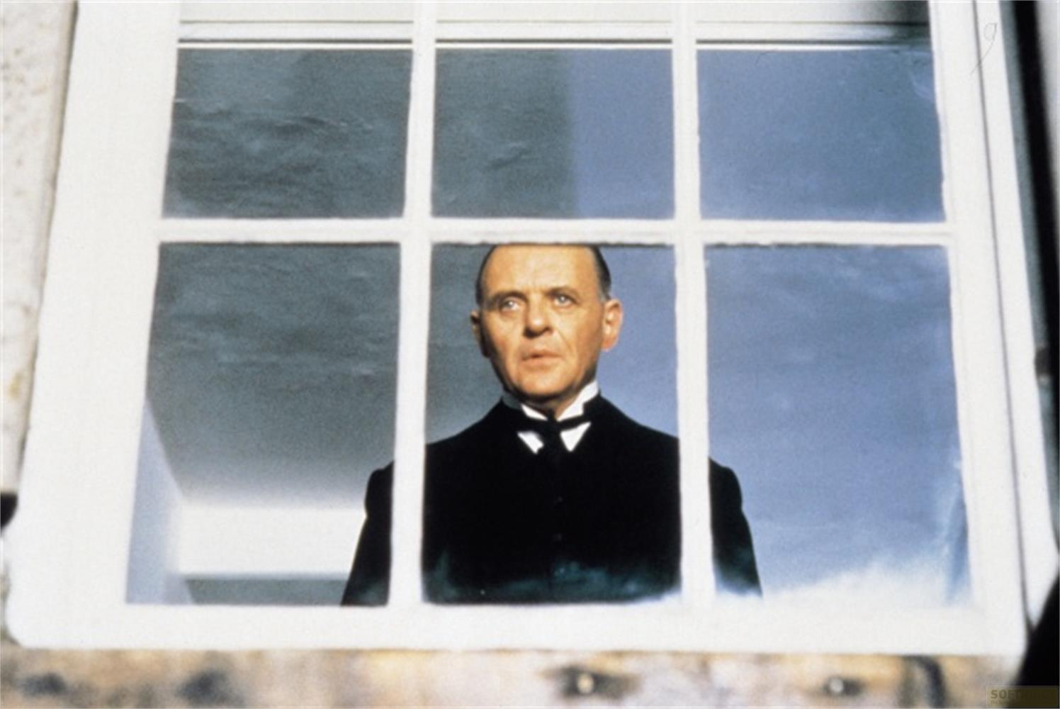 Ein Mann im schwarzen Anzug mit Fliege blickt durch ein Fenster.
