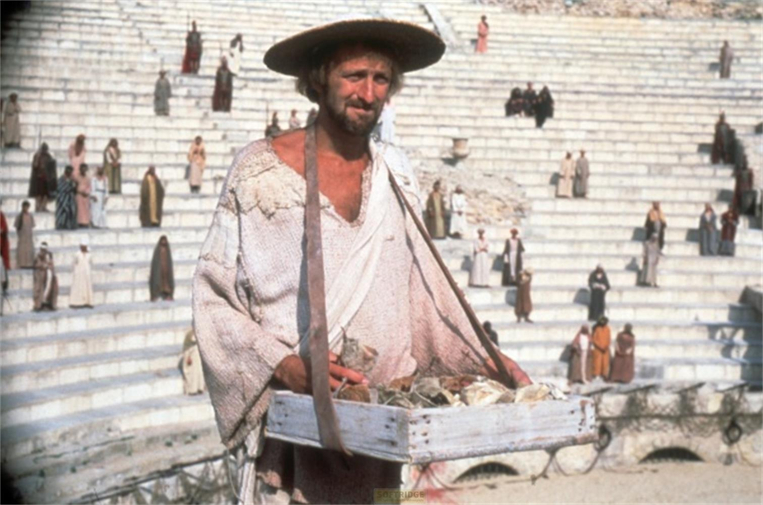 Un homme avec un chapeau et des vêtements en lambeaux devant une foule en gradins.