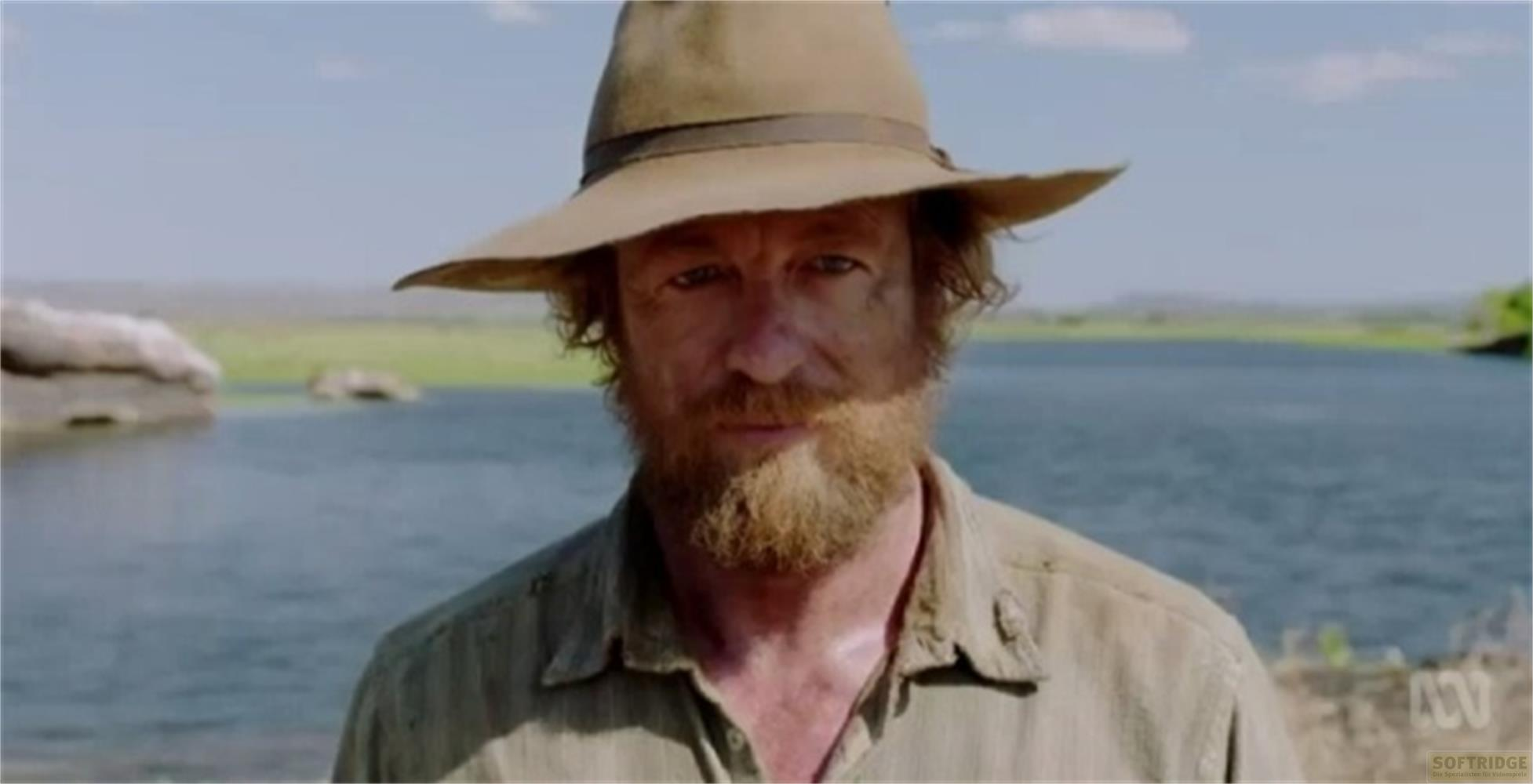 Un uomo con barba e cappello si trova davanti a uno specchio d'acqua.