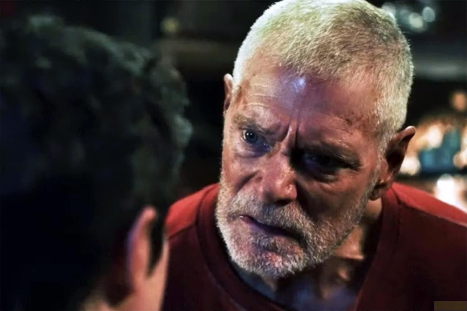 Un uomo anziano con capelli e barba grigi indossa una maglia rossa e appare preoccupato.