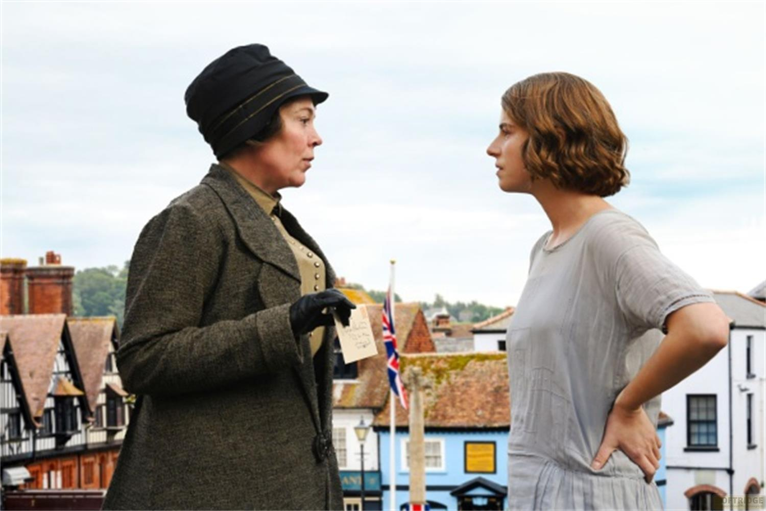Due donne conversano all'aperto con una scena cittadina di edifici e una bandiera sullo sfondo.