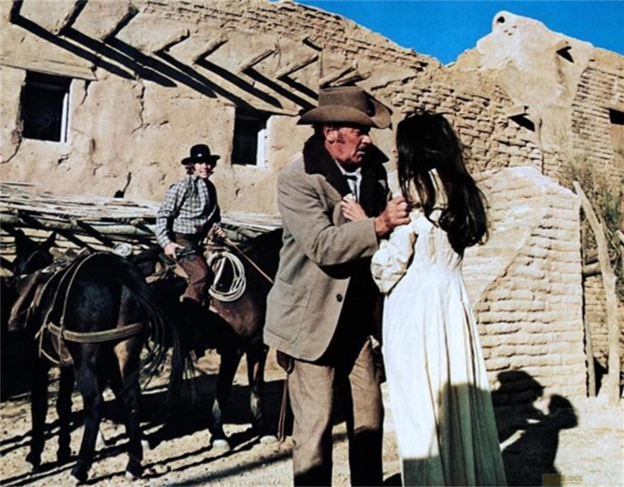 Uomo con cappello da cowboy parla con donna in vestito bianco. Ragazzo a cavallo.