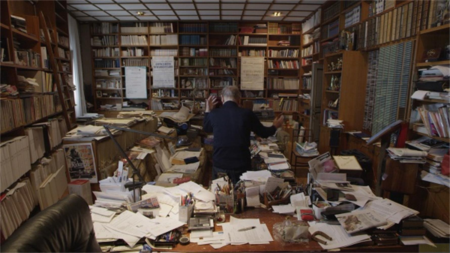 Uomo con camicia scura in stanza disordinata piena di libri e carte.