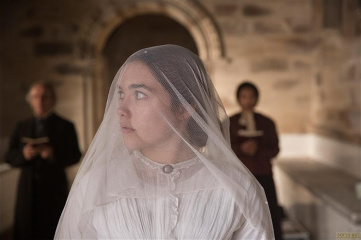 Una donna in abito bianco da sposa con velo guarda di lato. Due uomini sono visibili sullo sfondo.