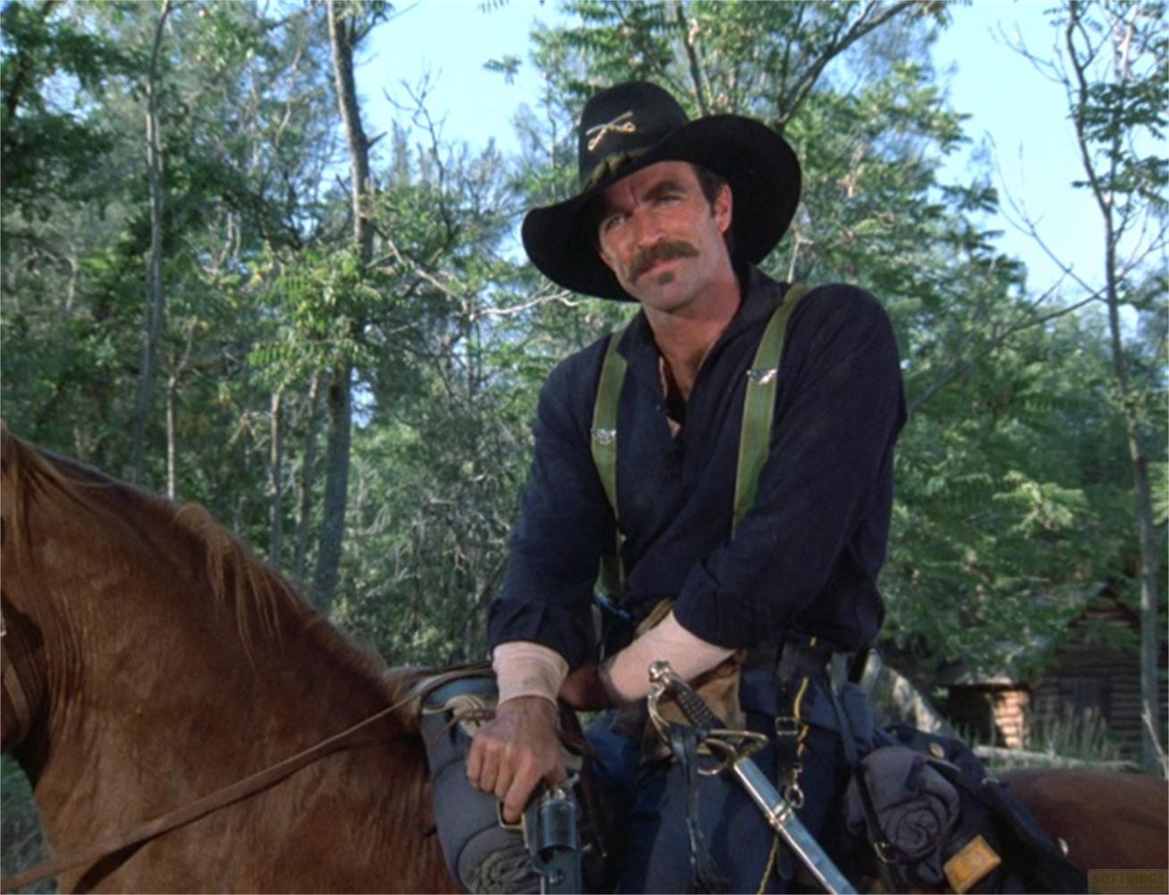 Uomo in uniforme con cappello e baffi cavalca un cavallo marrone nei boschi.