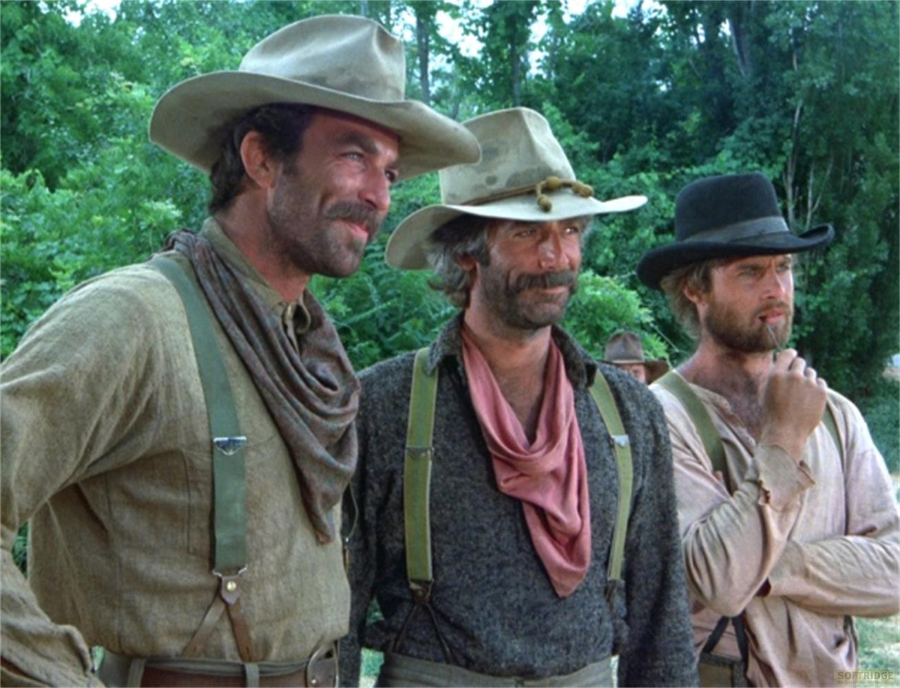 Tre uomini in abiti da cowboy, due in primo piano, con cappelli e bretelle.