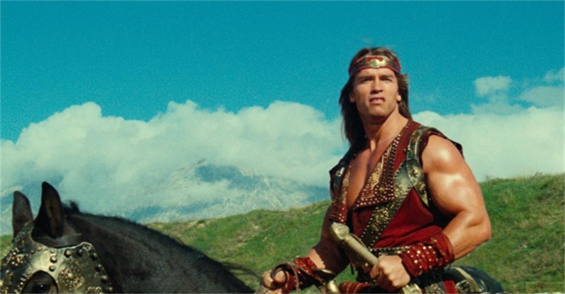 Arnold Schwarzenegger nei panni di Conan a cavallo con spada, contro cielo azzurro e montagne.