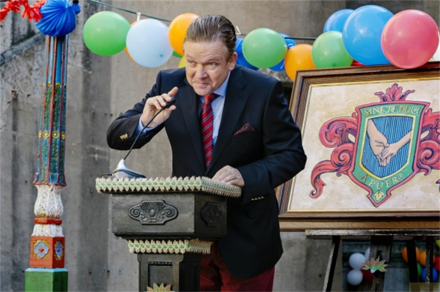 Ein Mann im dunklen Anzug spricht an einem Rednerpult. Ballons und ein Wappen sind im Hintergrund.