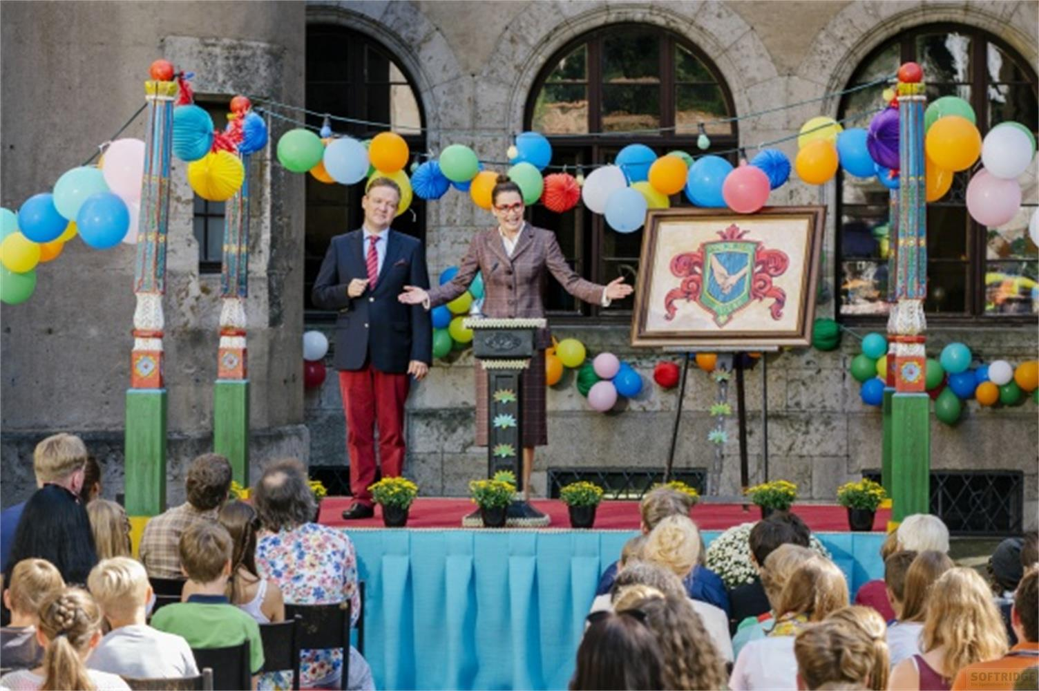 Zwei Personen stehen auf einer Bühne, flankiert von bunten Ballons und Dekorationen. Ein Publikum sitzt davor.