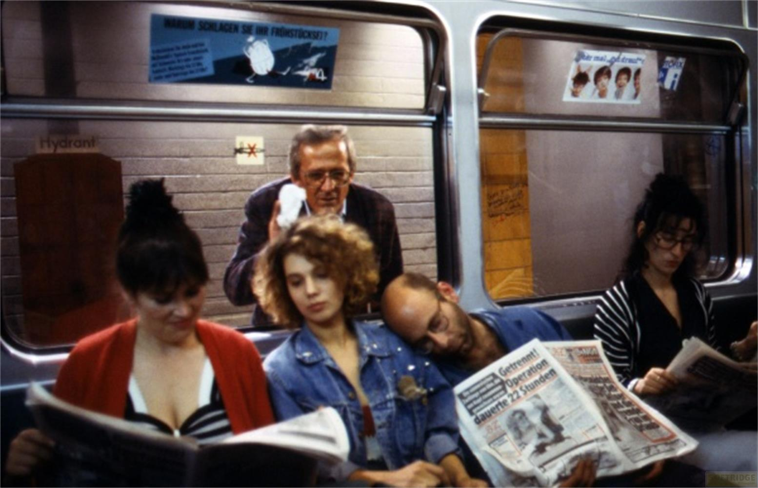 Persone su un treno leggono giornali, uno dorme. Un uomo si asciuga la fronte.