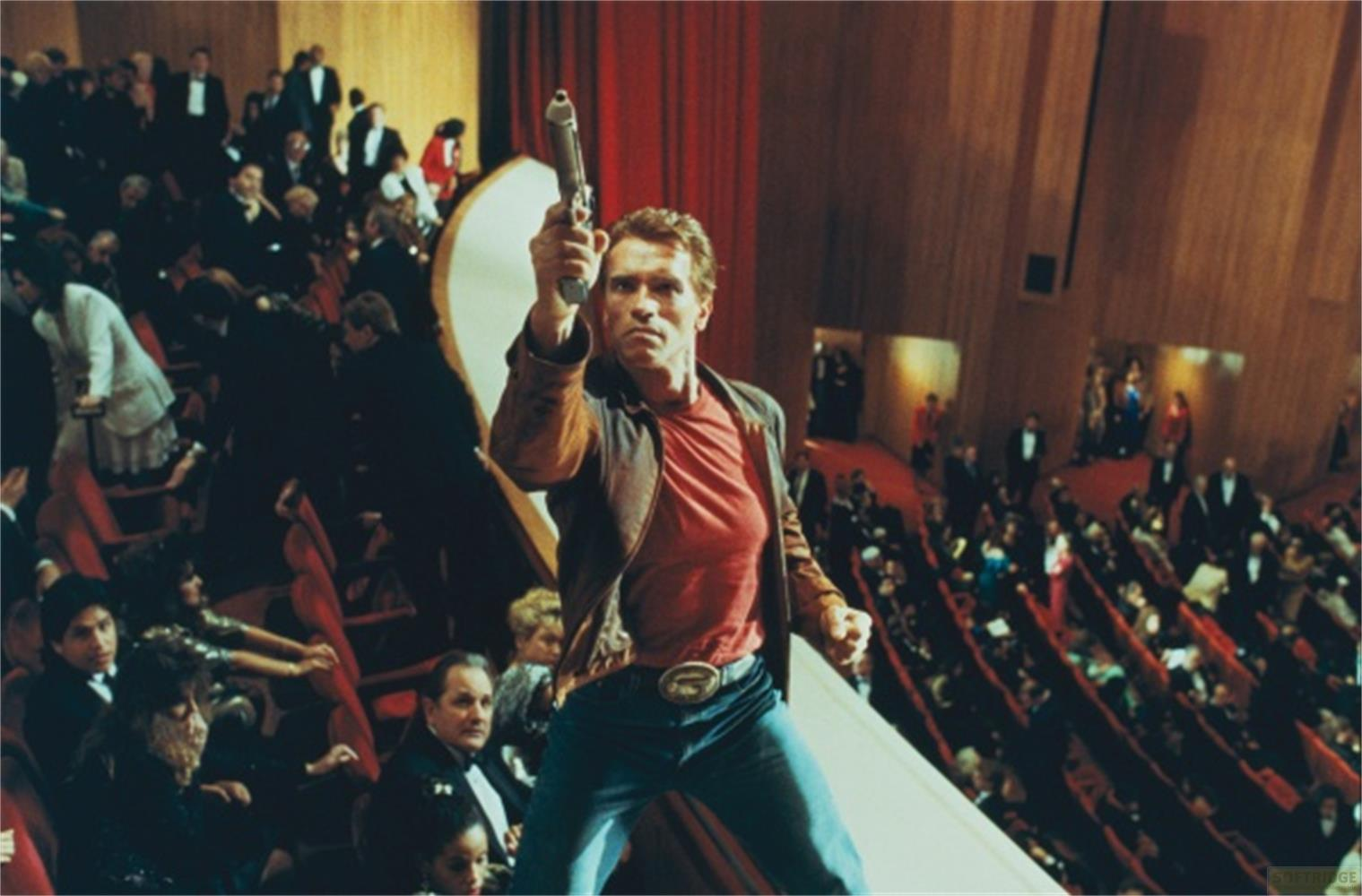 Uomo con pistola in un auditorium pieno con sedili rossi.