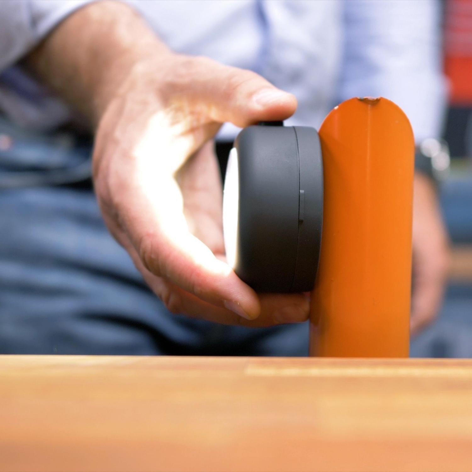 Una mano sostiene un dispositivo negro con luz brillante contra un poste naranja.